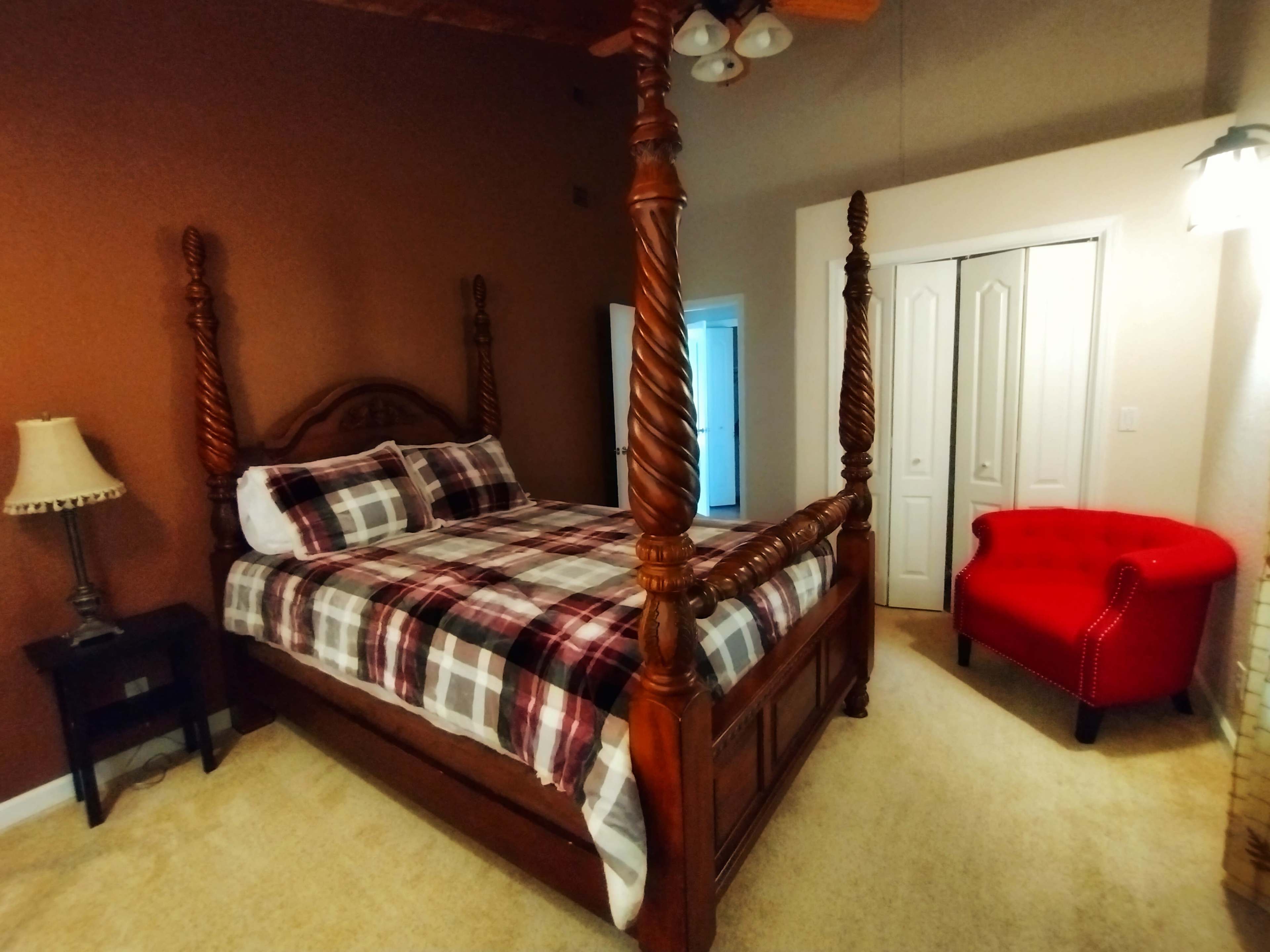 A bedroom featuring a four-poster wooden bed with a plaid comforter, a red accent chair, and a lamp on a nightstand.