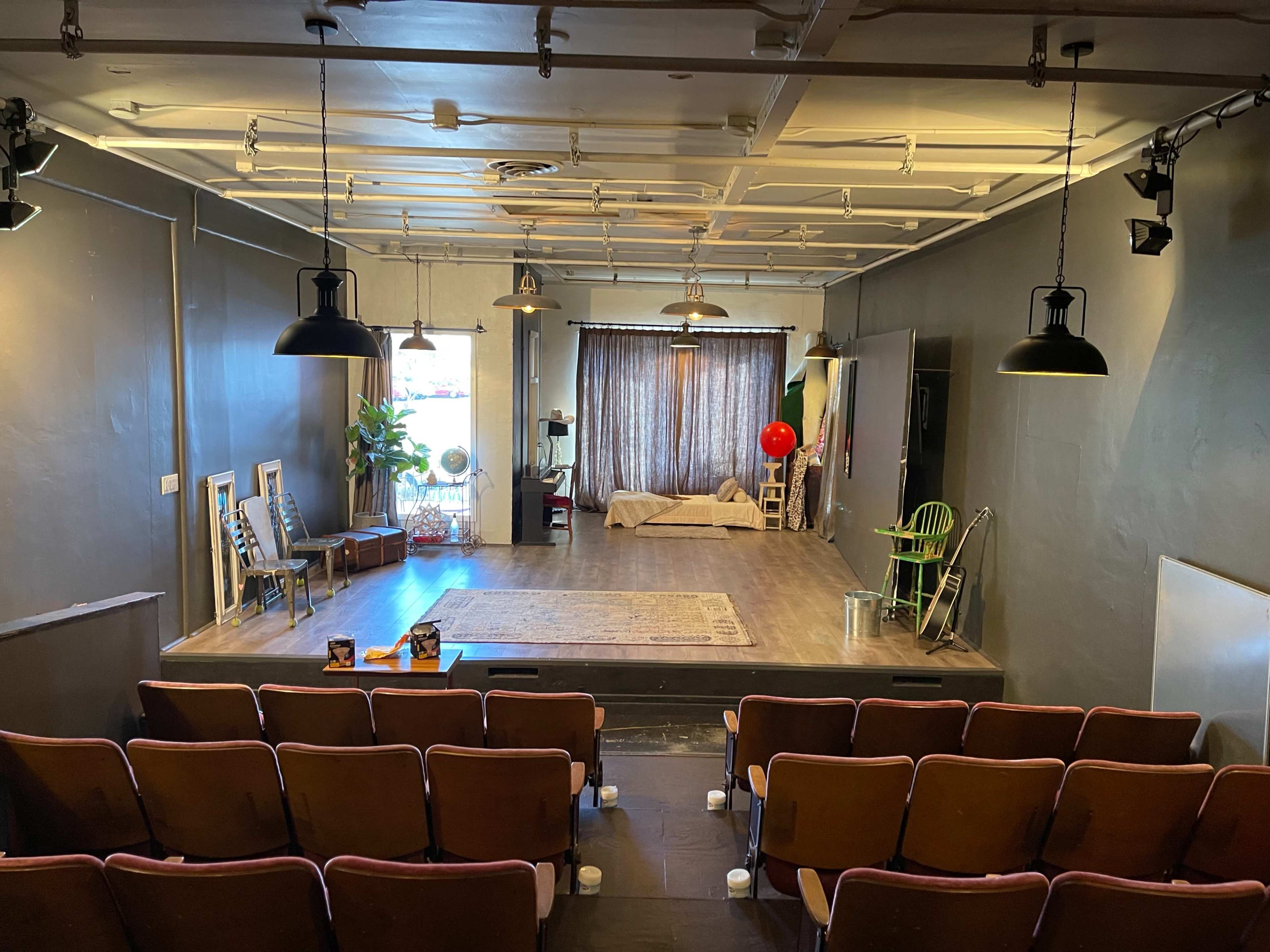 A theater stage with a minimalist set, including a large curtain, a bed, a green chair, and several potted plants, viewed from a seating area with reddish-brown chairs.