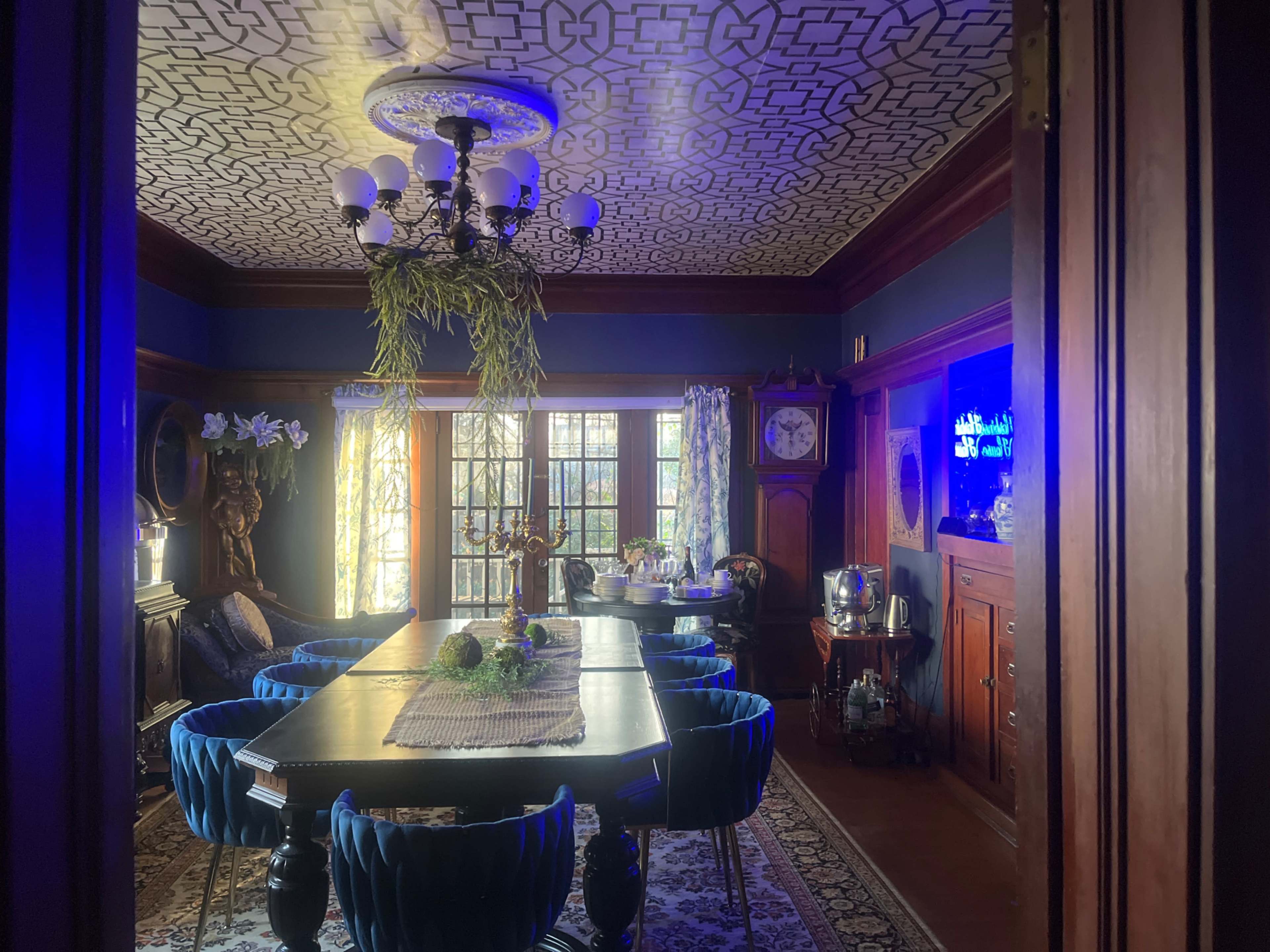 The image shows a dining room with a patterned ceiling, a long table surrounded by blue chairs, and large windows letting in natural light.