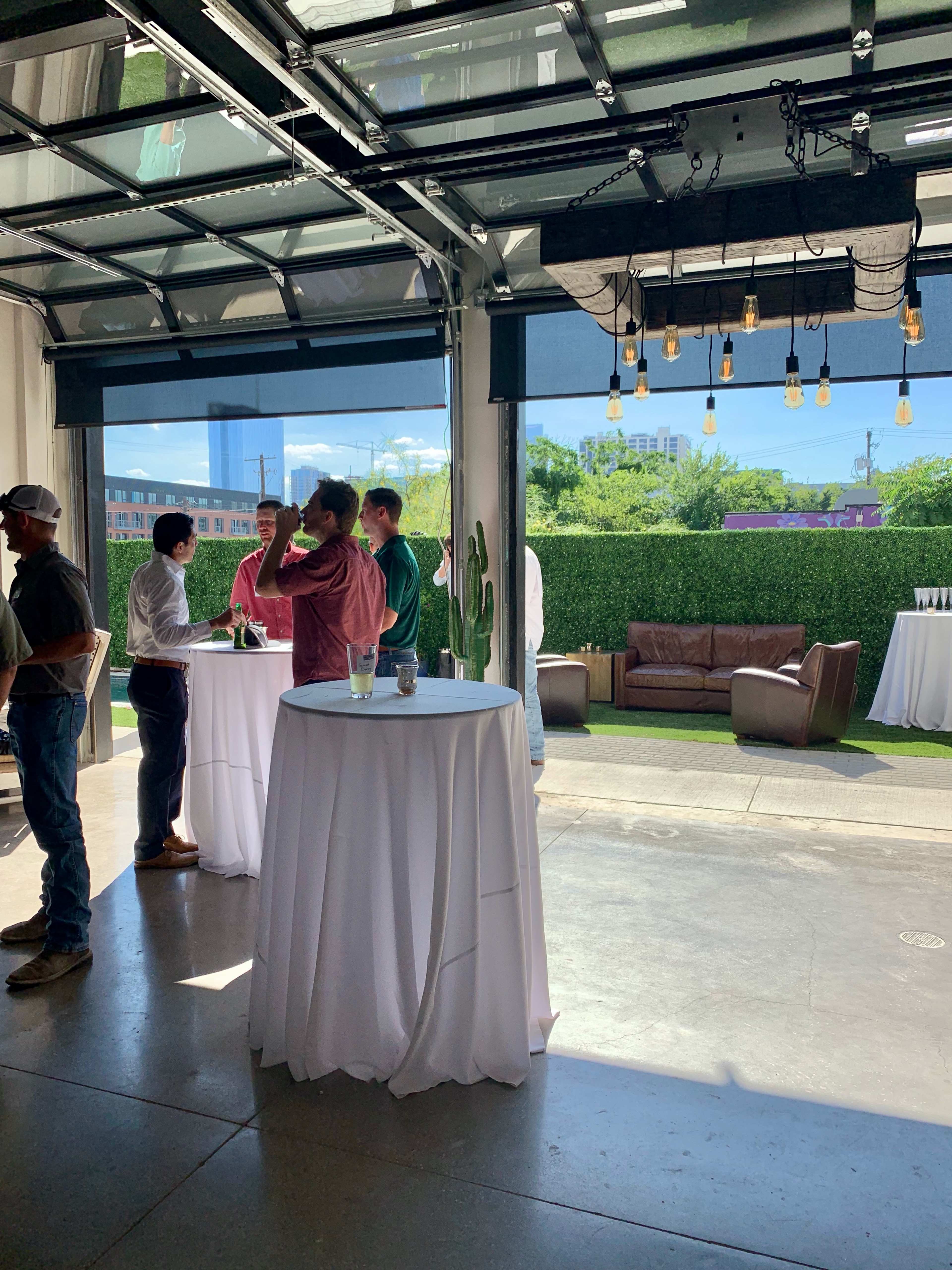 A group of people are socializing at high-top tables in a brightly lit indoor-outdoor event space with greenery in the background.