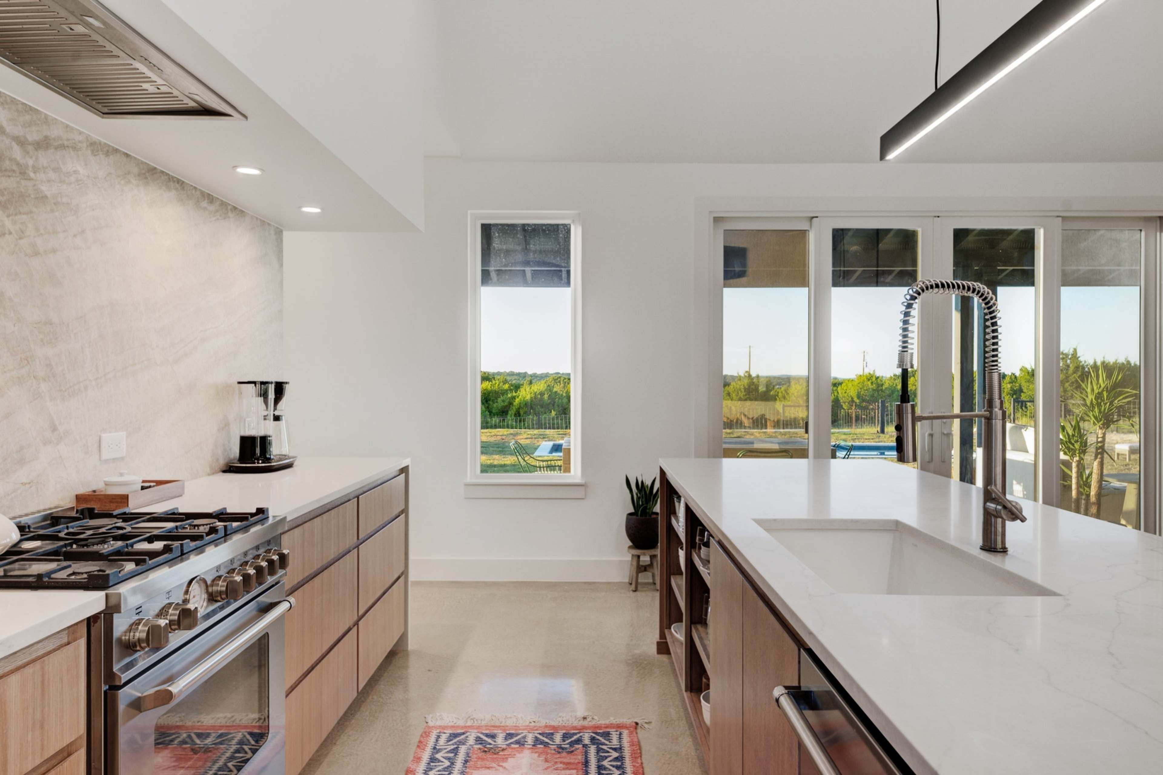 A modern kitchen features a stainless steel gas stove, a central island with a sink, and large windows overlooking a pool and greenery.