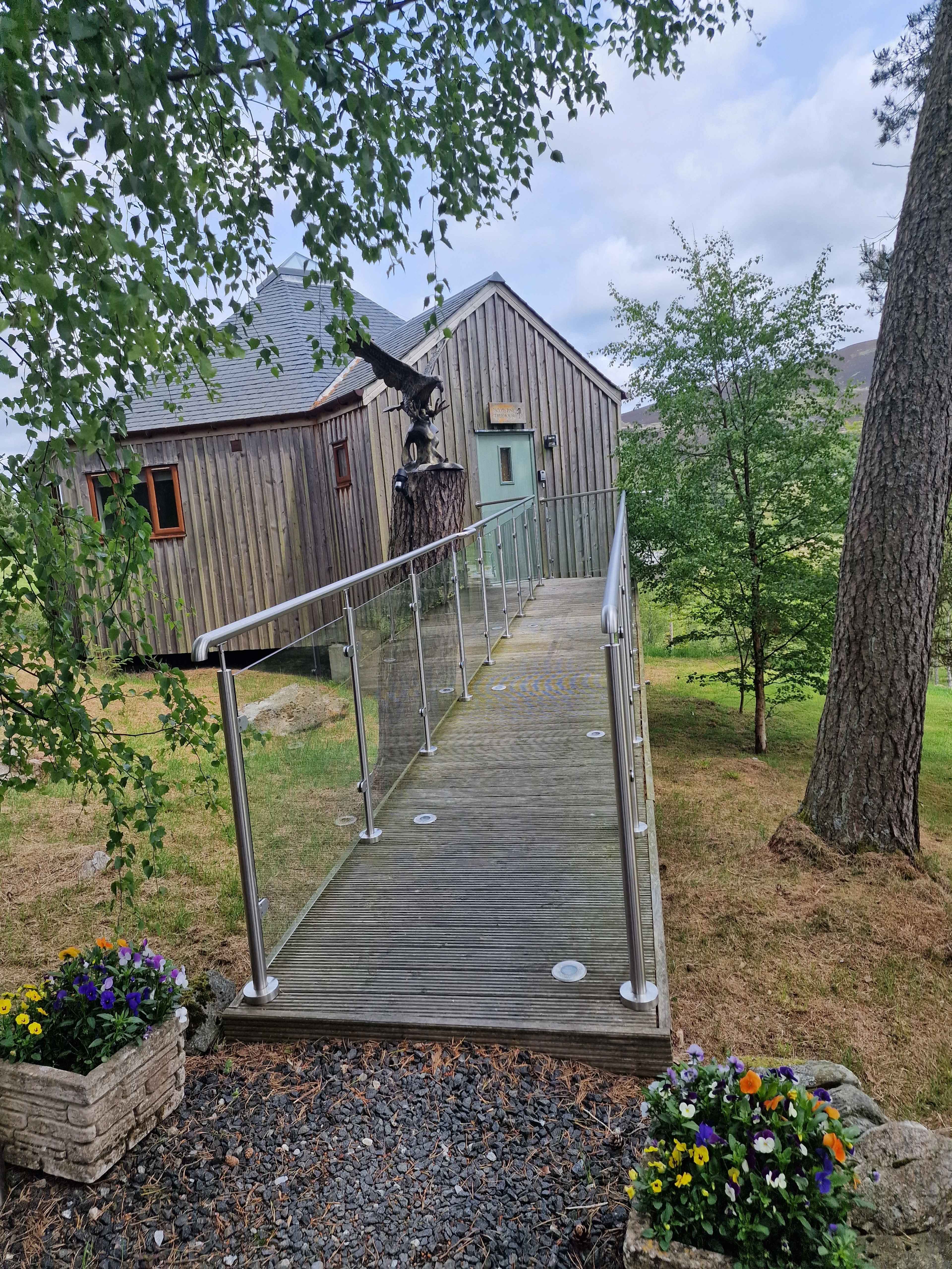 A wooden ramp with glass railings leads to a modern wooden building surrounded by trees and flower planters.