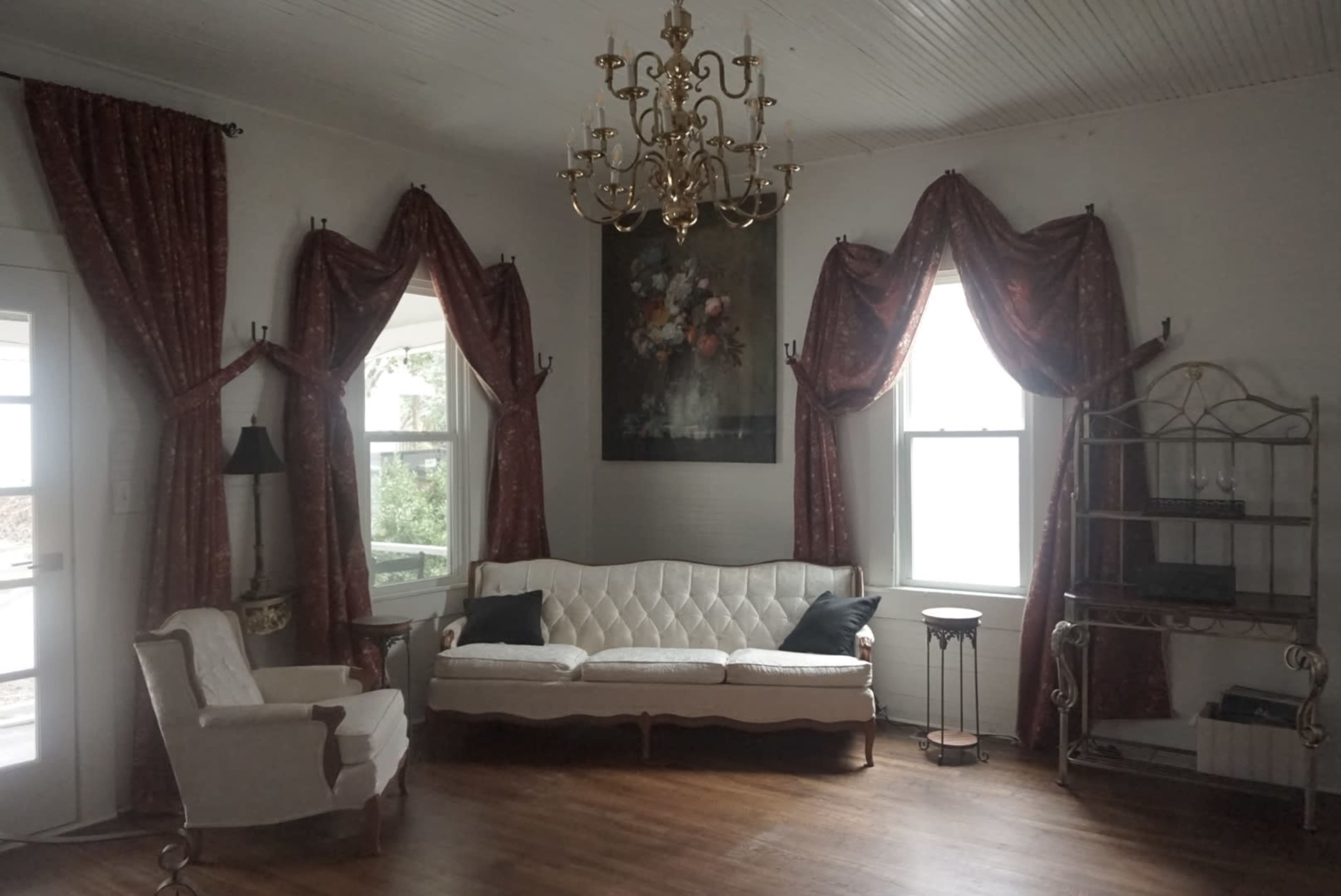 A cozy living room with a white upholstered sofa, two decorative windows with curtains, and a chandelier hanging from the ceiling.