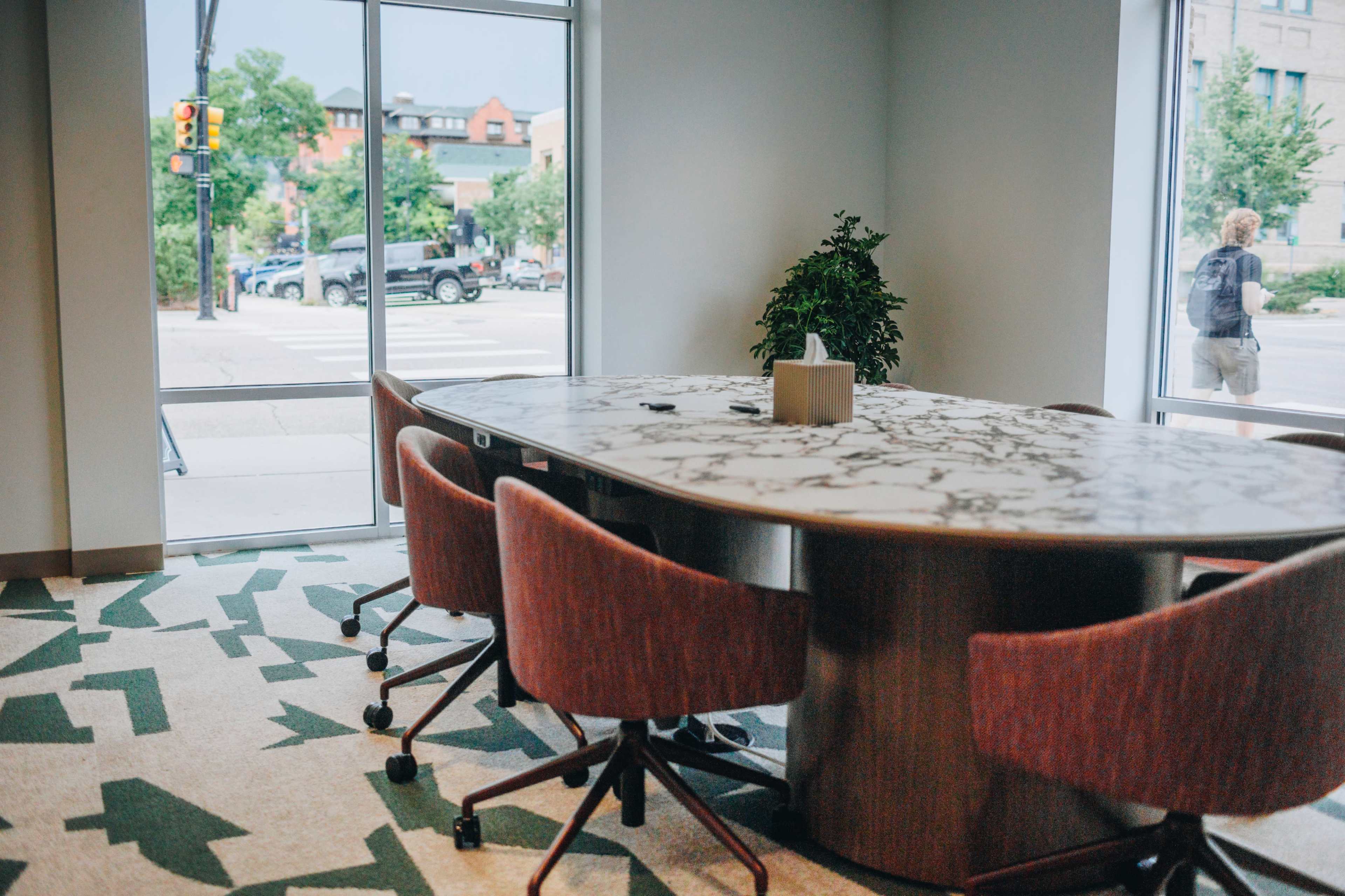 A modern conference room features a large marble table surrounded by upholstered chairs, with windows overlooking a street corner and greenery outside.