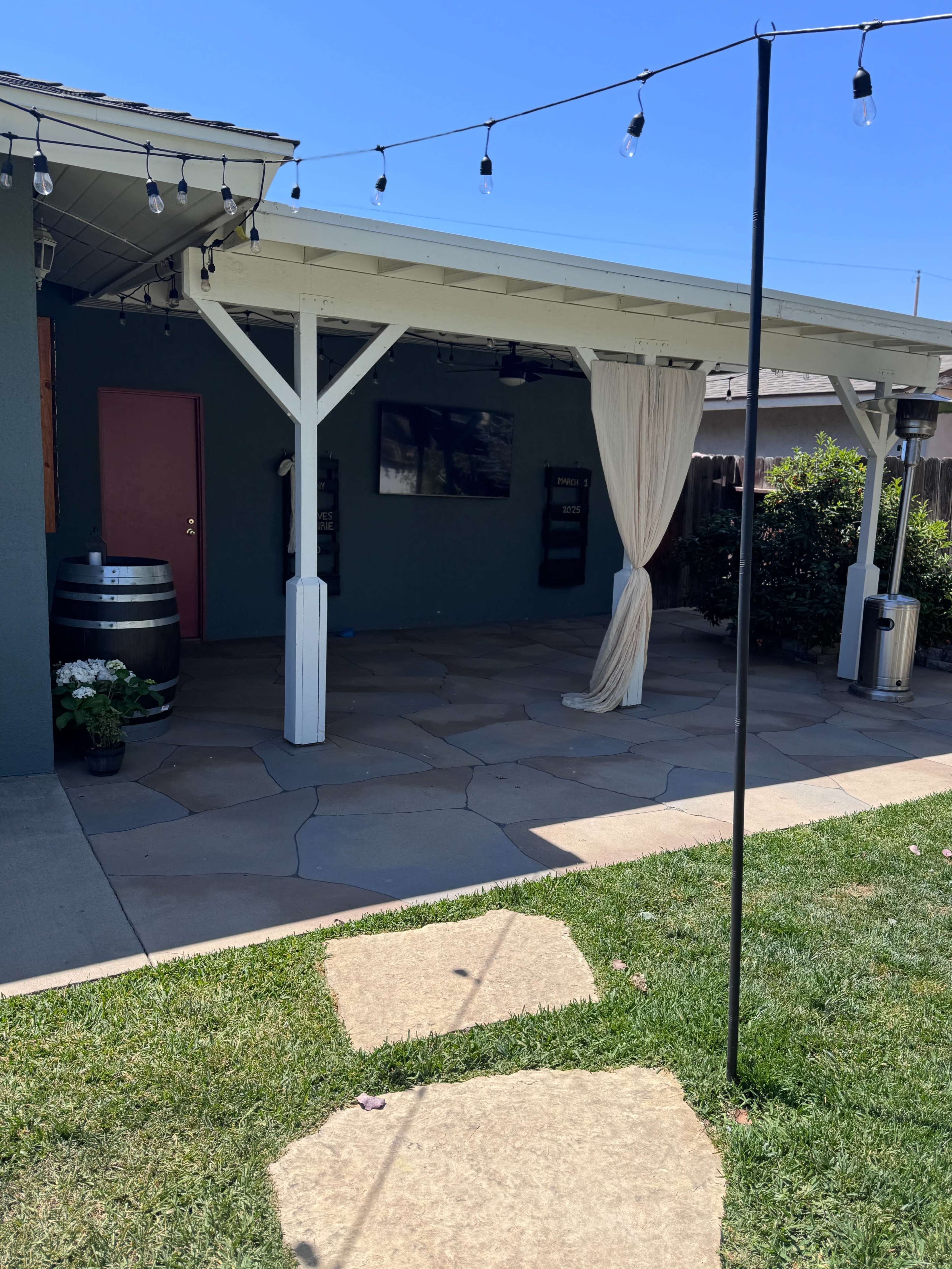 The image shows a patio area featuring a covered space with a decorative curtain, a barrel, and string lights, surrounded by grass and concrete pathways.