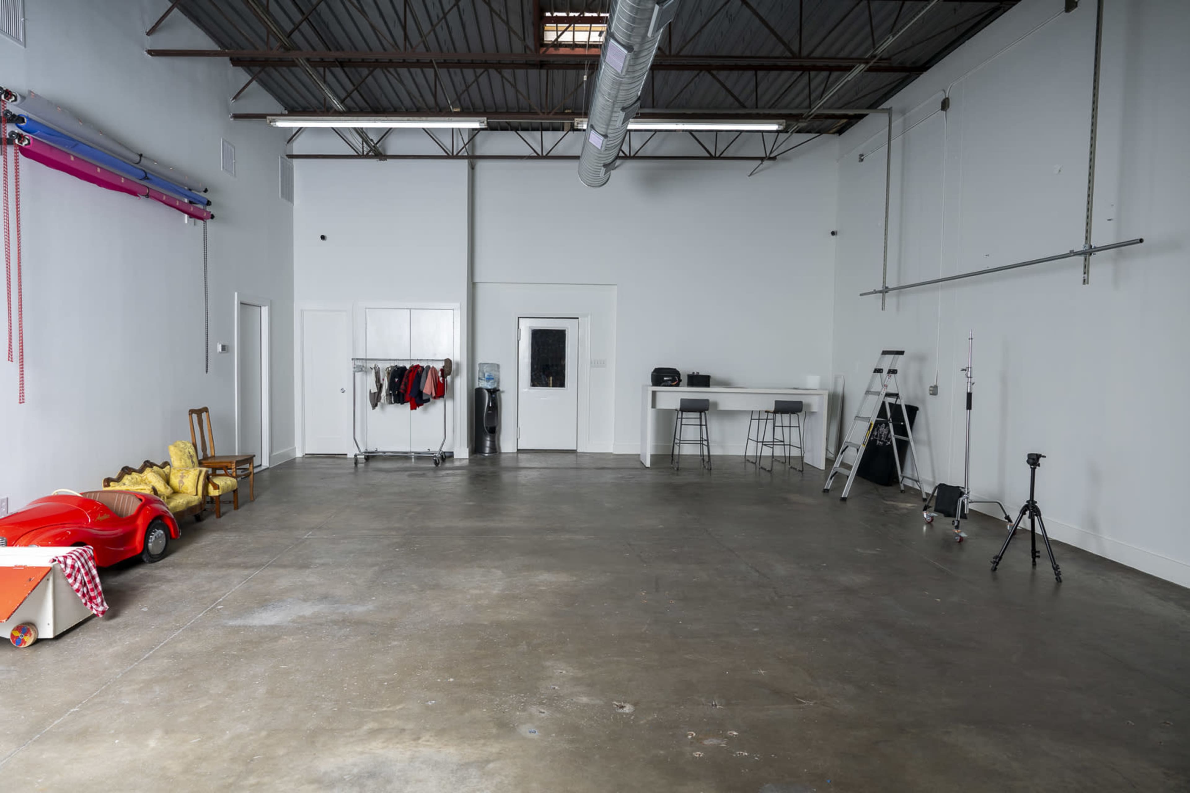 The image shows a spacious, empty garage-like room with concrete flooring, a bar-height table and stools, and various props including a red toy car and a clothing rack.