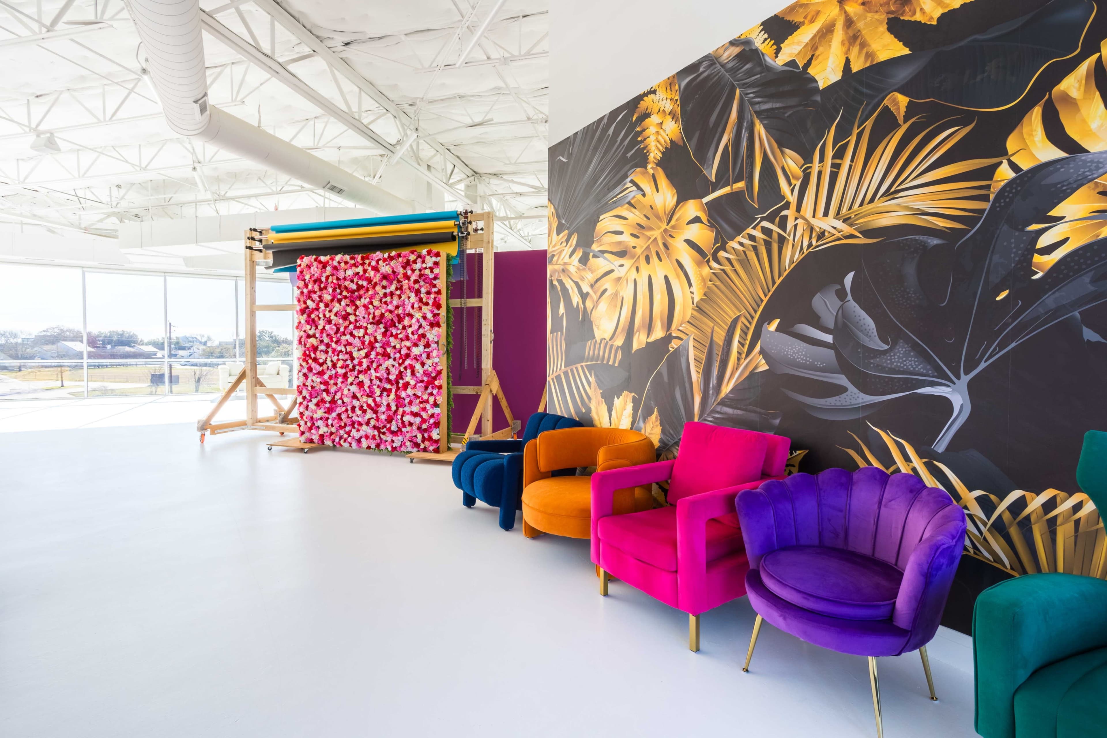 The image shows a brightly decorated interior space featuring colorful velvet chairs and a large wall mural of tropical leaves alongside a floral backdrop on a wooden frame.