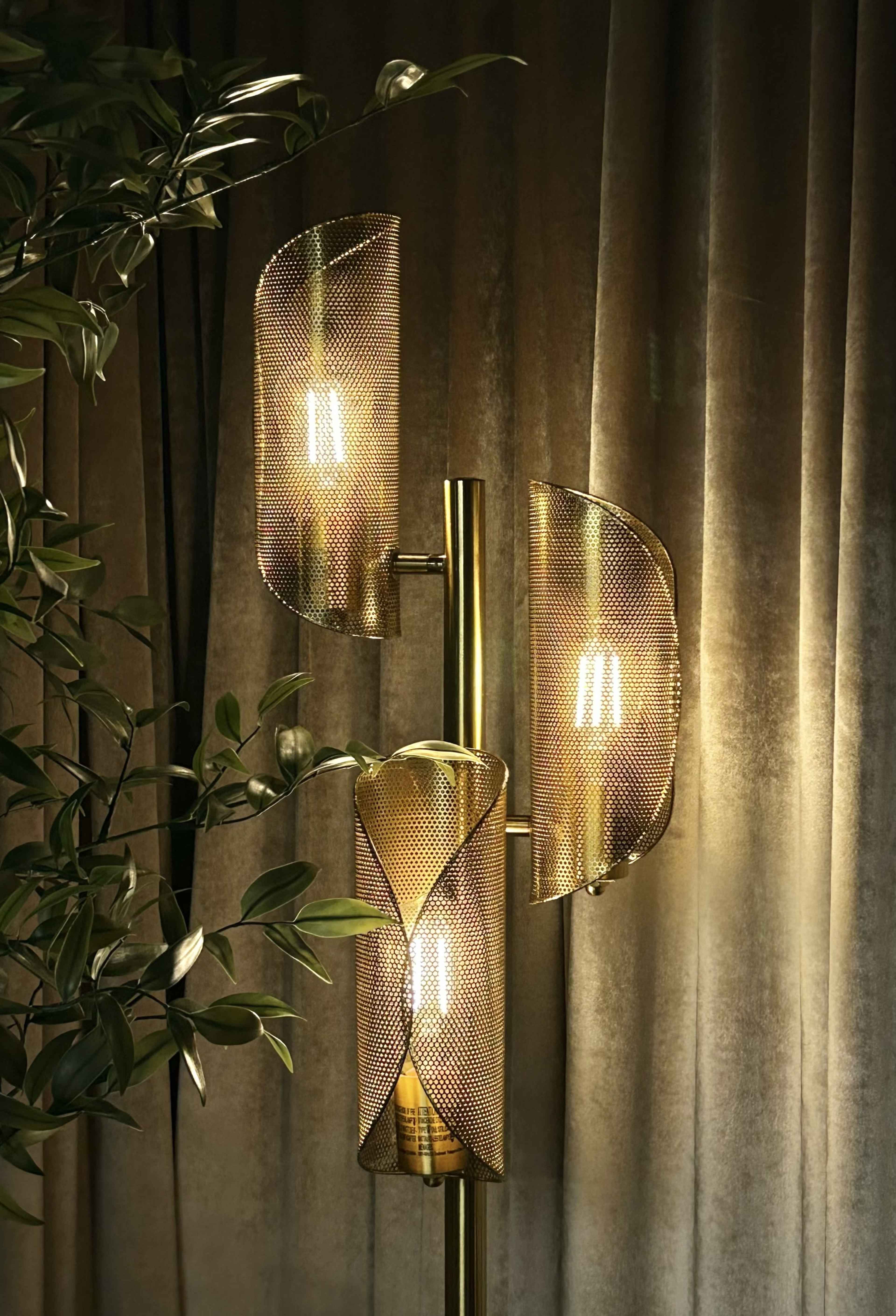 The image features a modern floor lamp with three mesh shade fixtures illuminated against a backdrop of dark, velvety curtains.