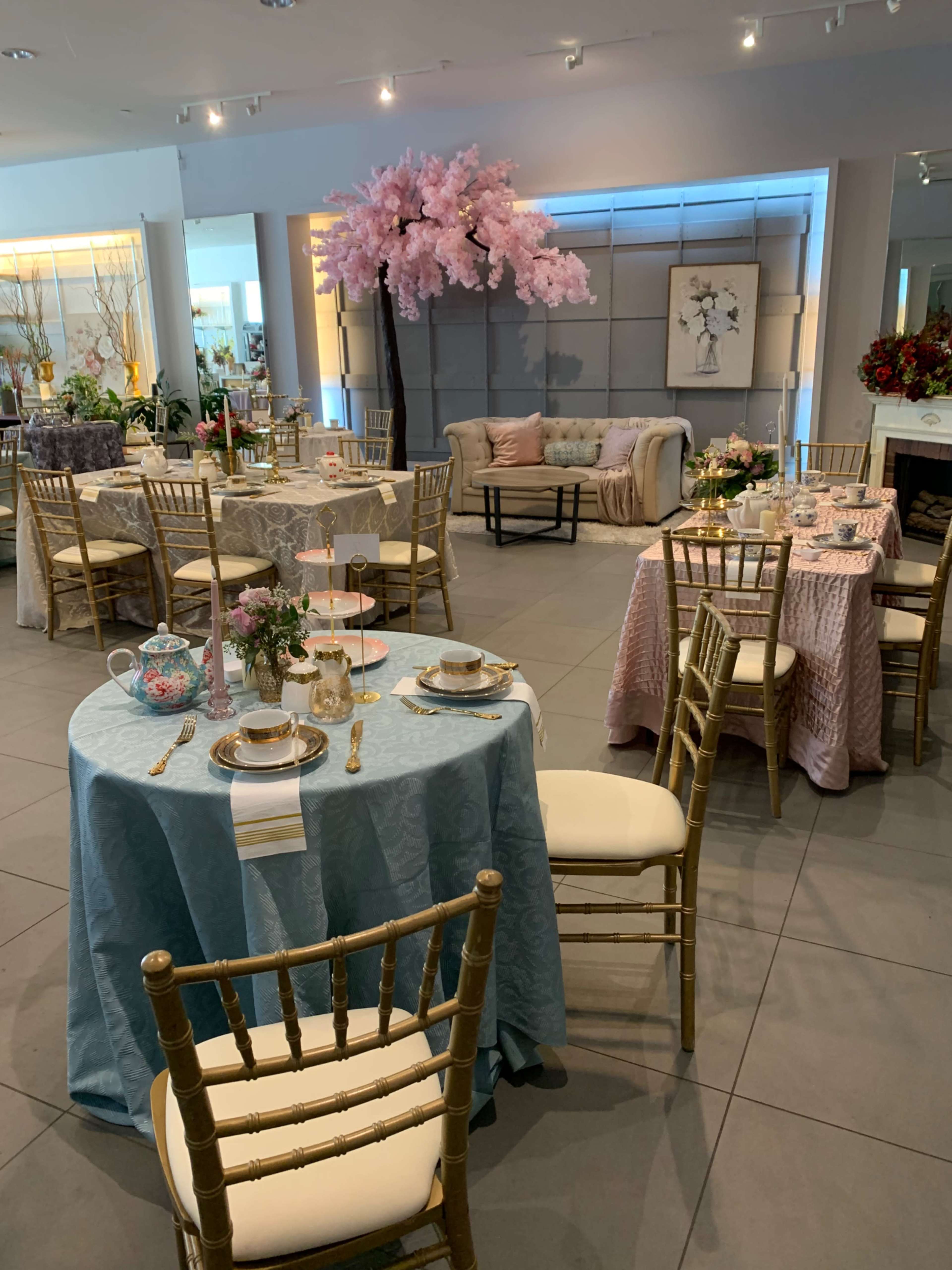 The image shows a beautifully arranged event space with elegantly decorated tables, featuring pastel-colored tablecloths, floral centerpieces, and a large cherry blossom display in the background.