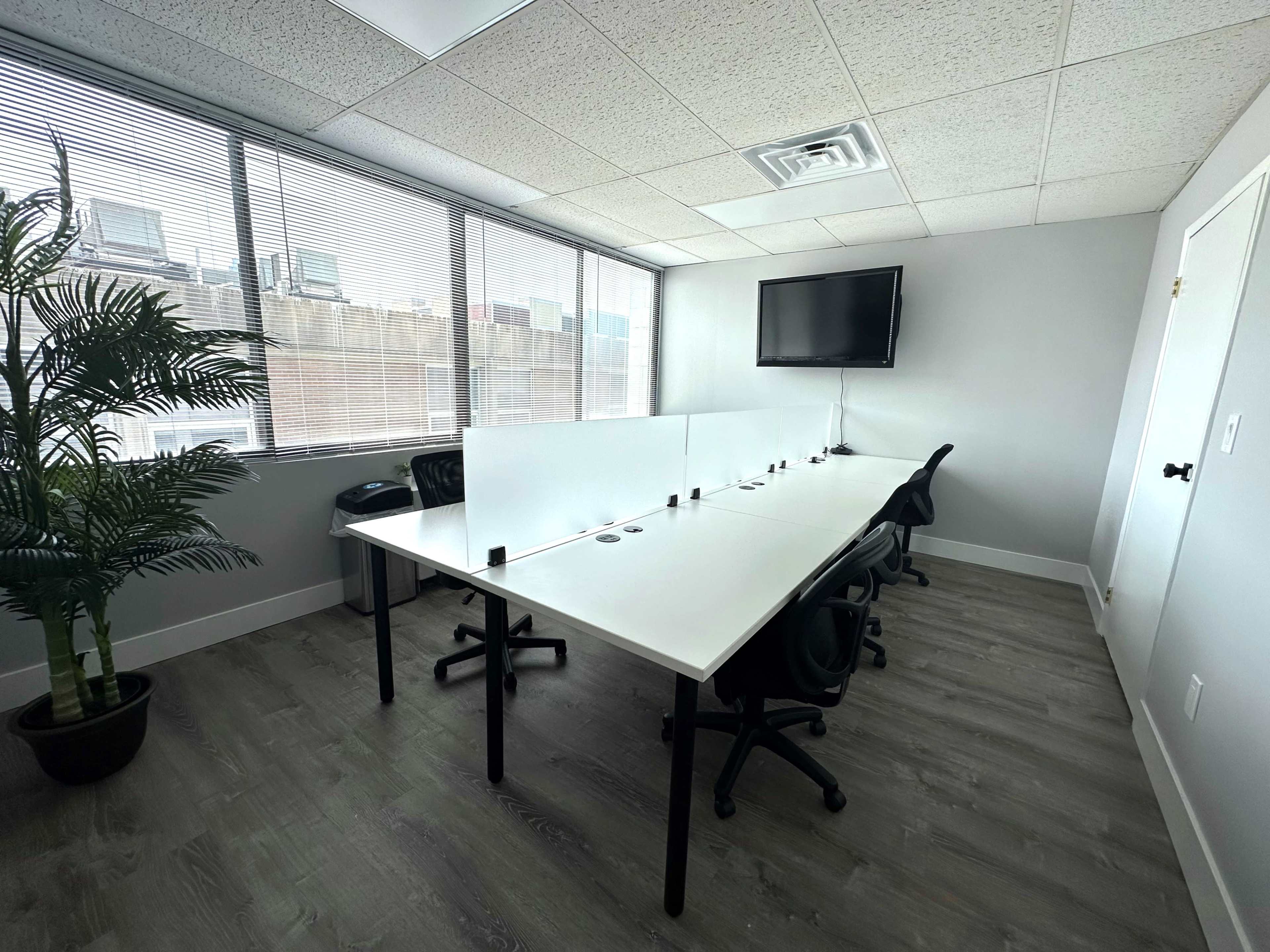 A modern conference room features a long white table with several black chairs and a wall-mounted television, illuminated by natural light from large windows.