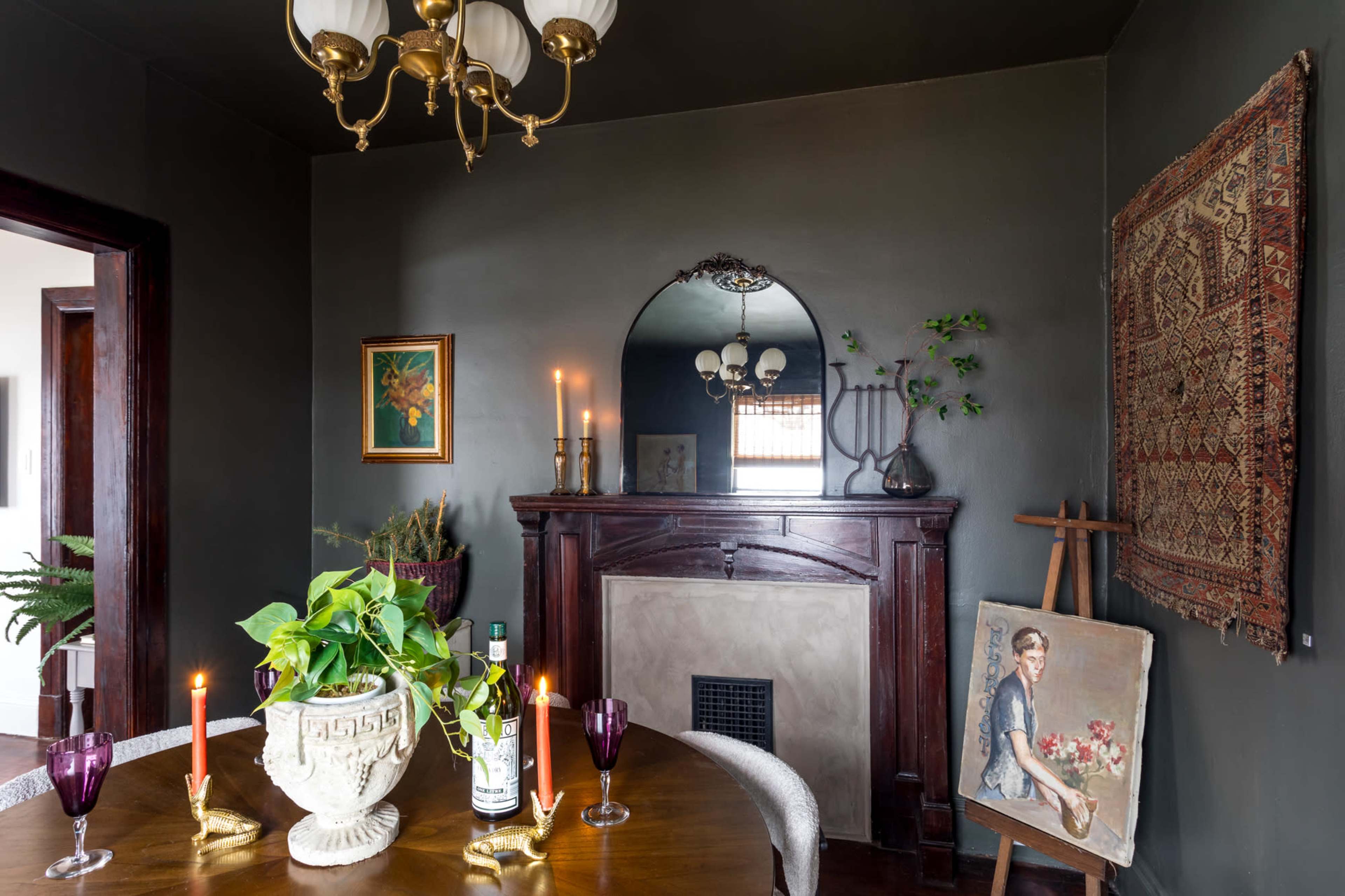 A dining area with a wooden table, a decorative fireplace, a framed painting, and a vase of greenery, illuminated by candles.