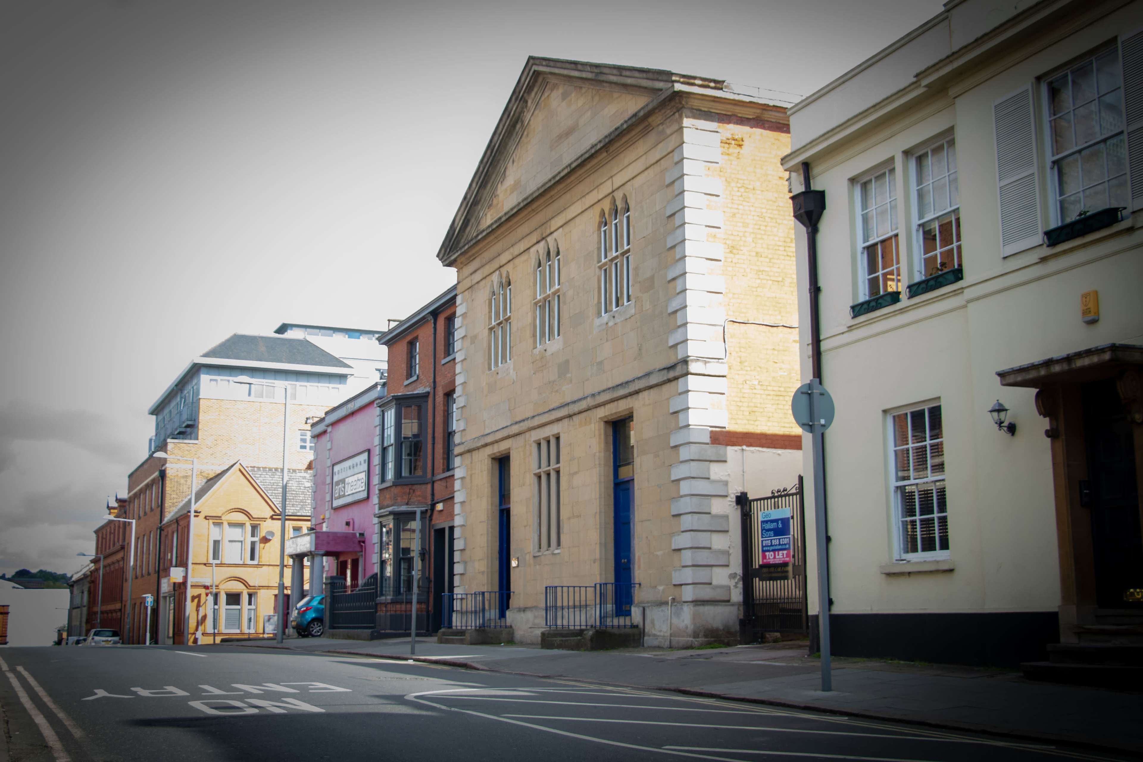 A street lined with buildings, featuring a prominent stone structure with a pediment on the left and more modern buildings on the right.