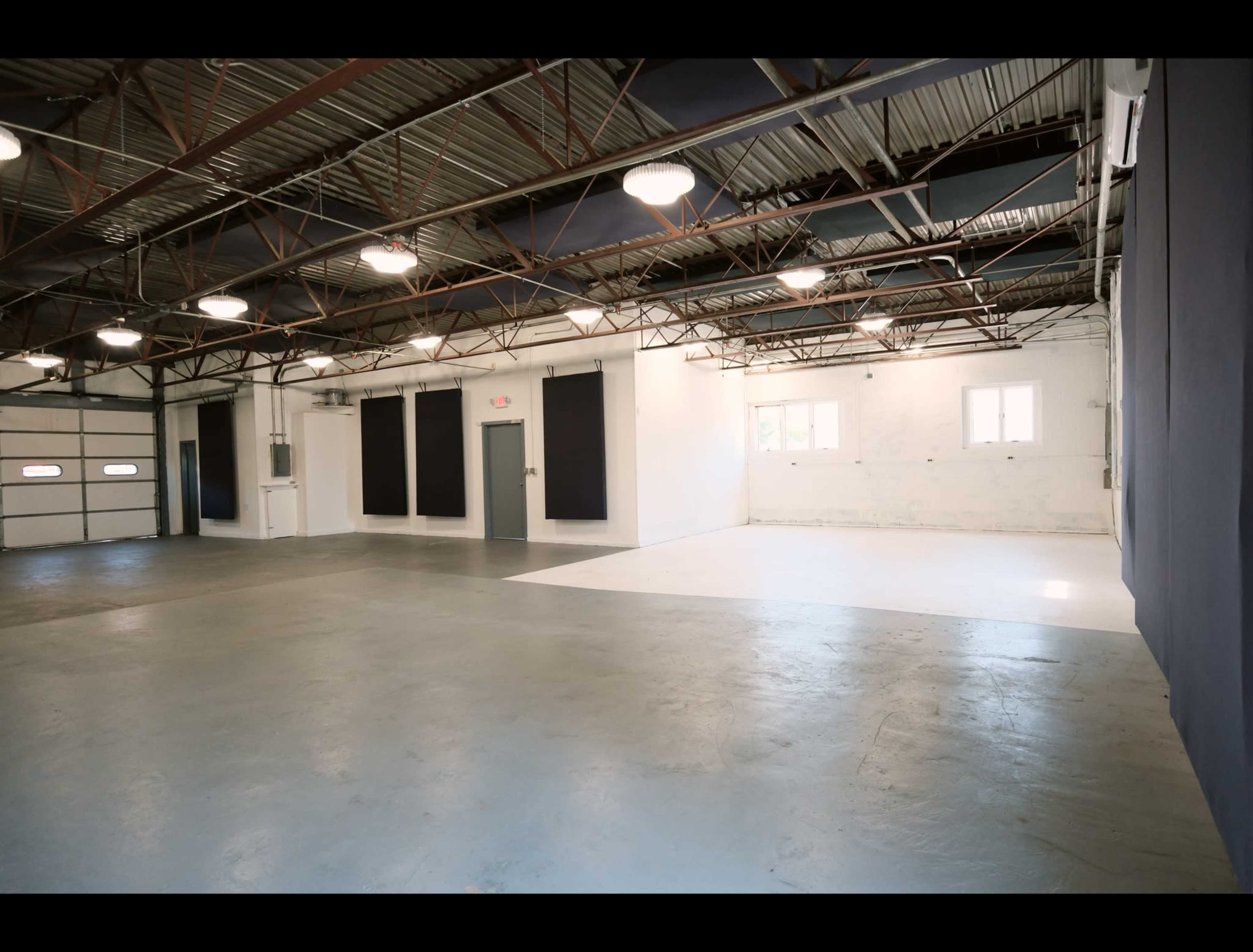 The image shows a spacious, industrial-style interior with concrete floors, exposed metal beams, and white walls, featuring large windows and a garage door.