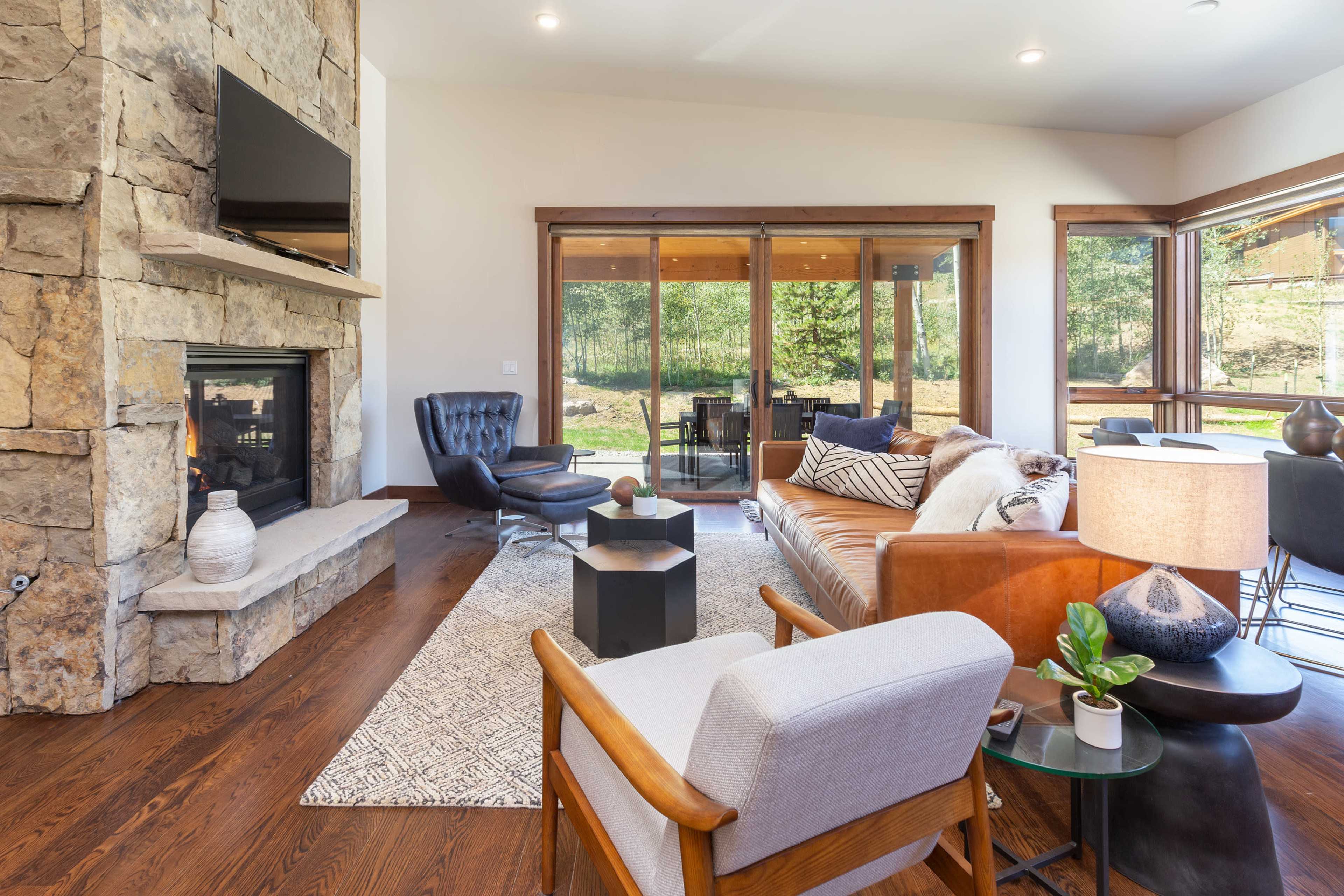 The living room features a stone fireplace, a leather sofa, and large windows that open to a view of the outdoor area.