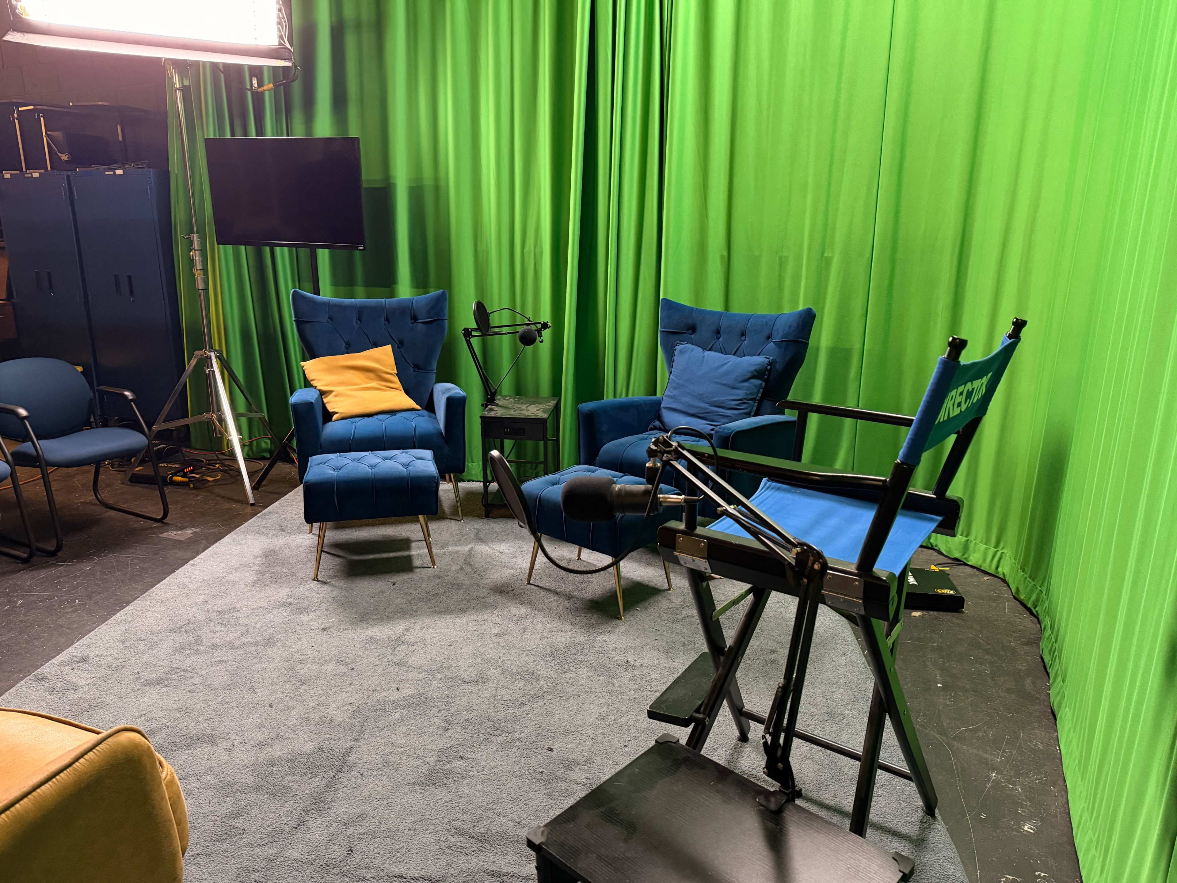 A recording studio features two blue chairs, a director's chair, and green curtains in the background.