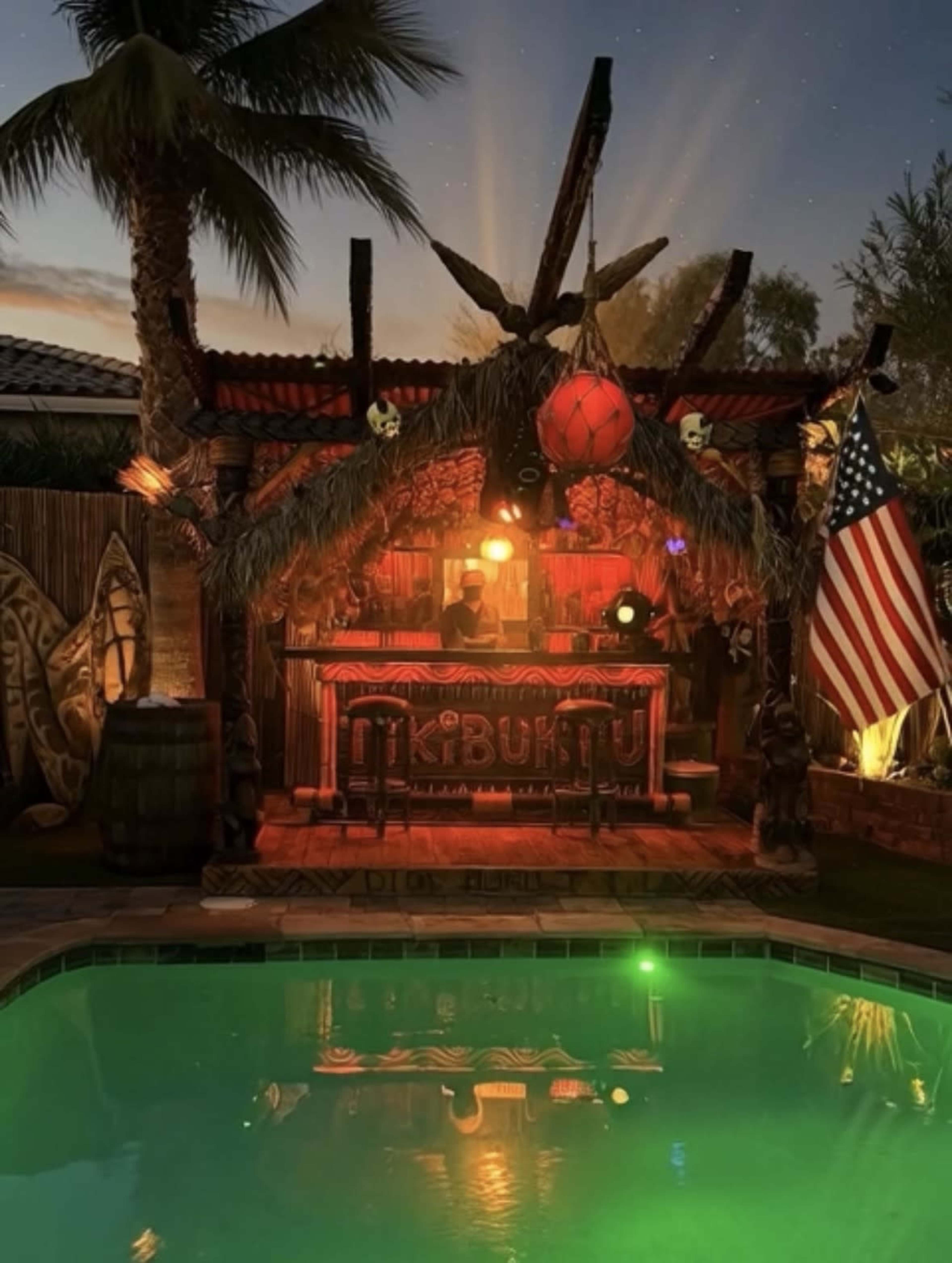 The image shows a tiki bar with vibrant lighting, surrounded by palm trees, a swimming pool, and an American flag displayed nearby during twilight.