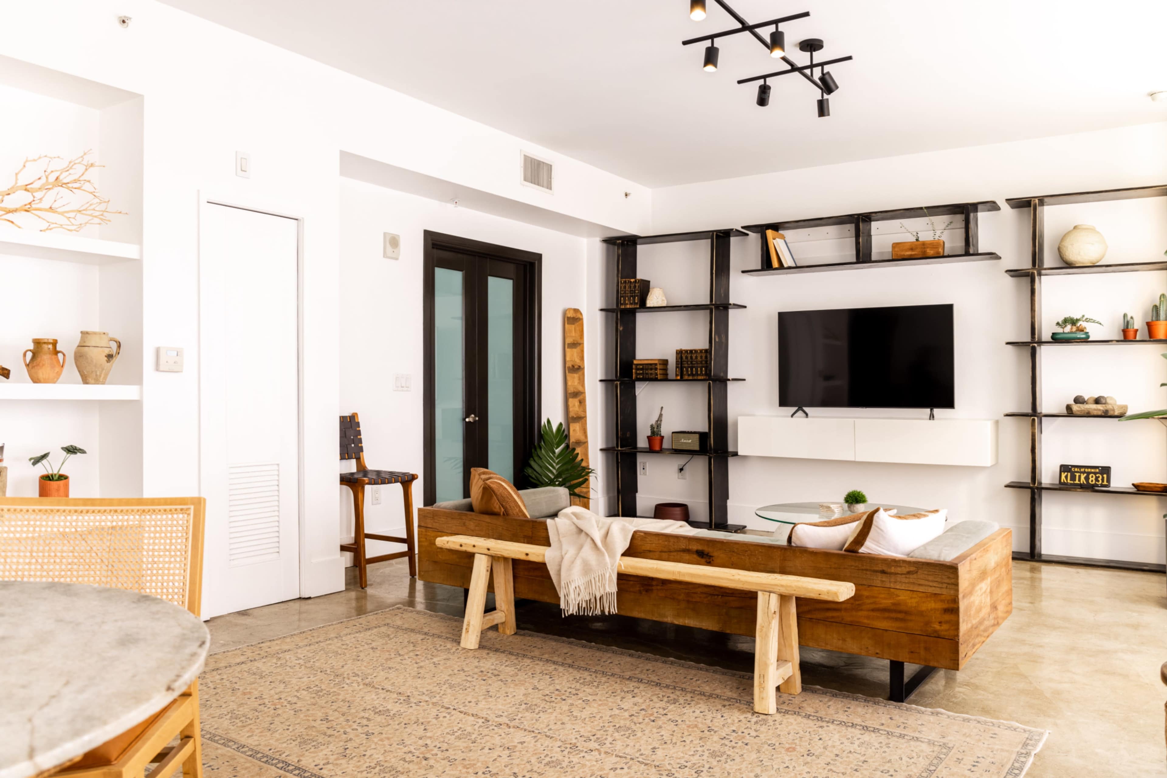 A modern living room with a wooden sofa, a coffee table, and a wall-mounted television, set against a backdrop of minimalist shelving filled with decorative items.