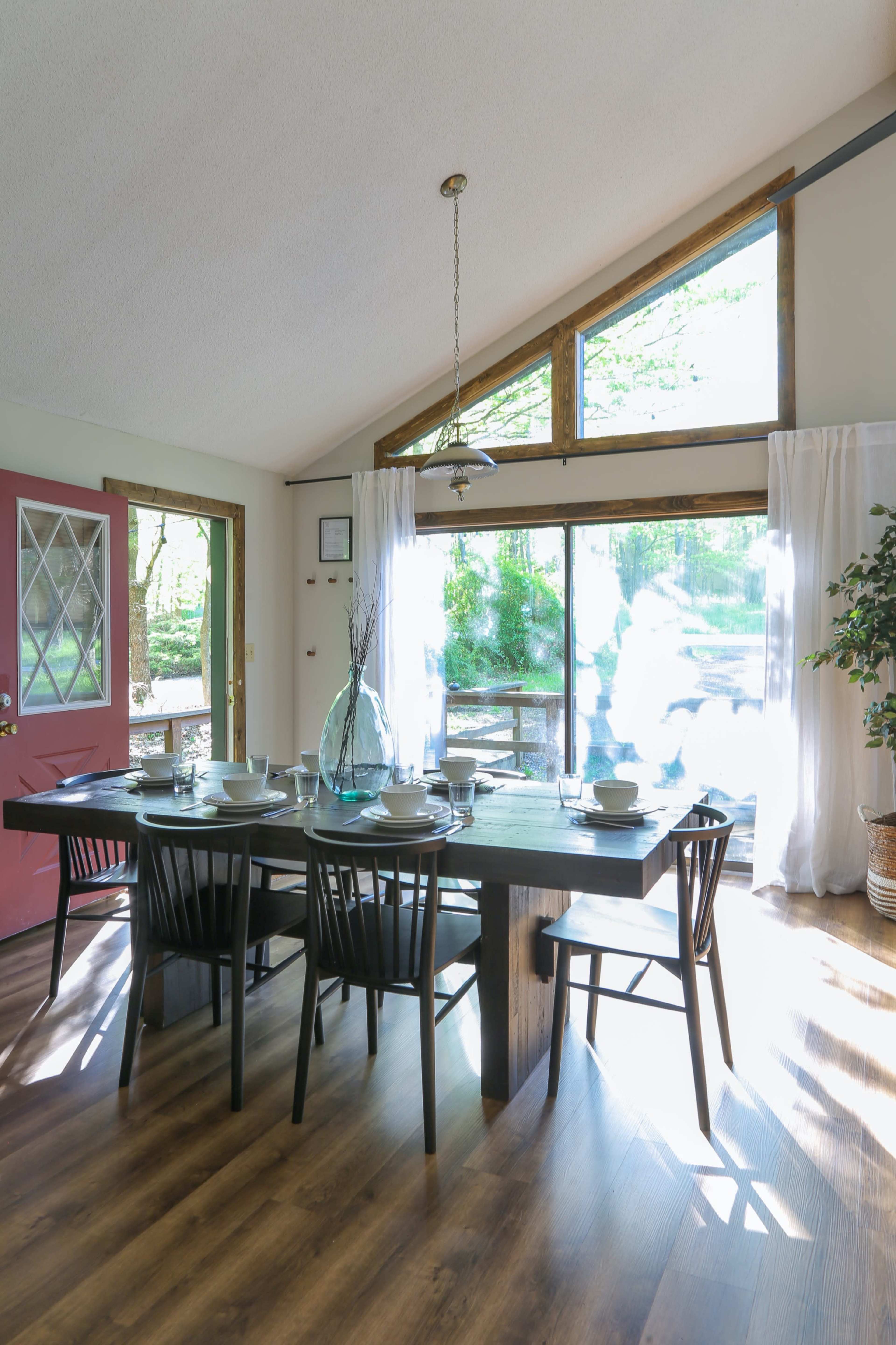 A dining area with a wooden table set for a meal, surrounded by black chairs, and large windows allowing natural light to fill the space.