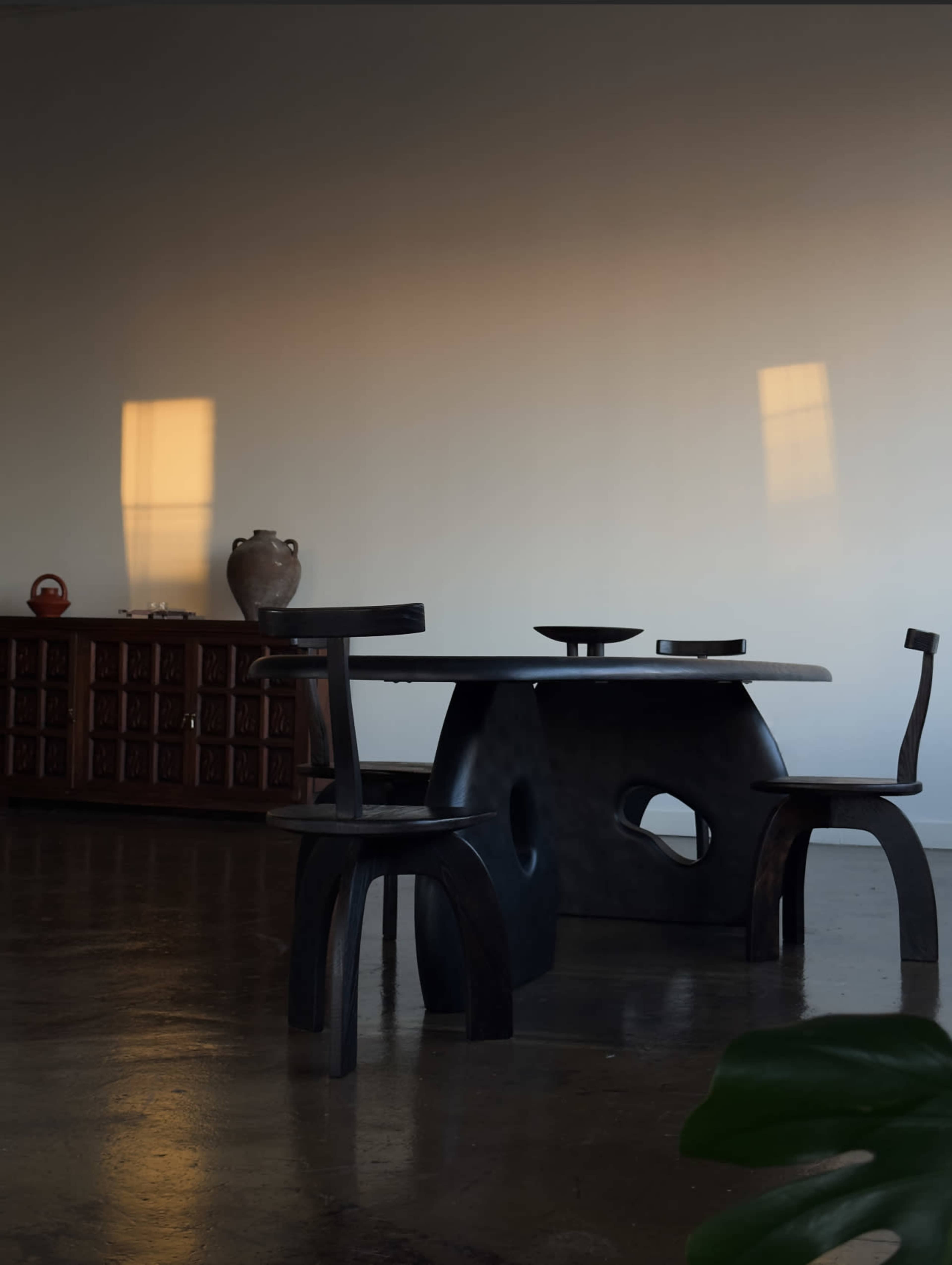 A dark, modern dining table with two matching chairs sits in a minimalist room, with a wooden cabinet and a vase in the background.