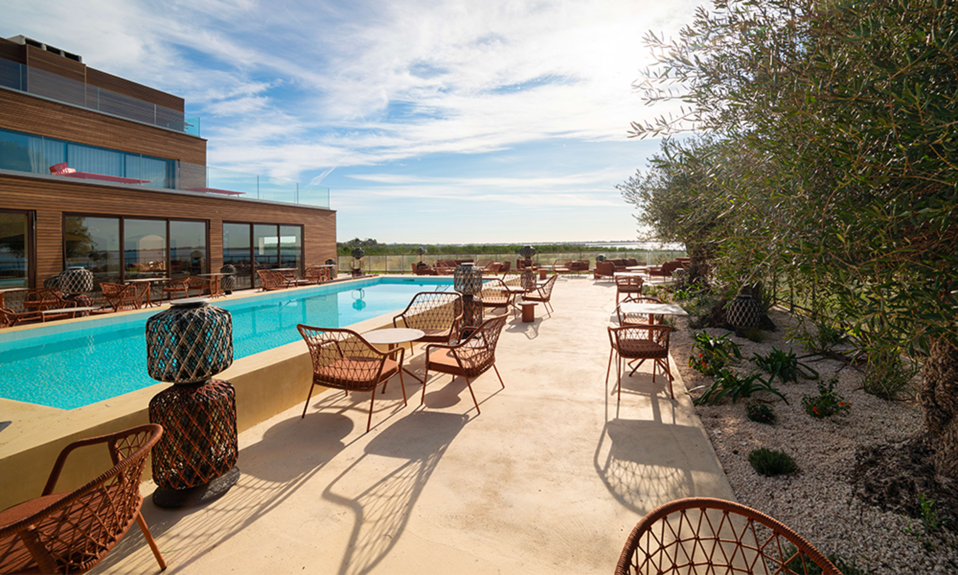 The image shows a poolside area with woven chairs and tables, surrounded by greenery and a clear sky.