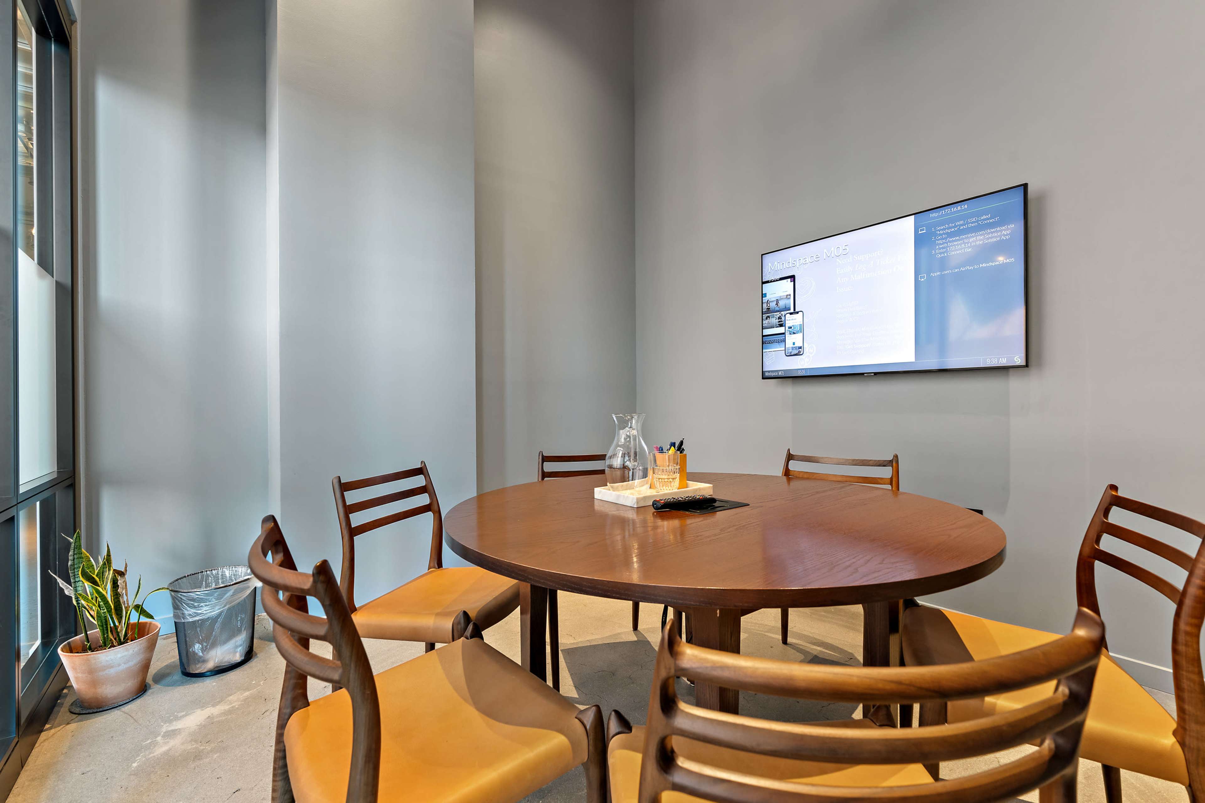 A round wooden table surrounded by six chairs is positioned in a meeting room with a wall-mounted screen displaying information.