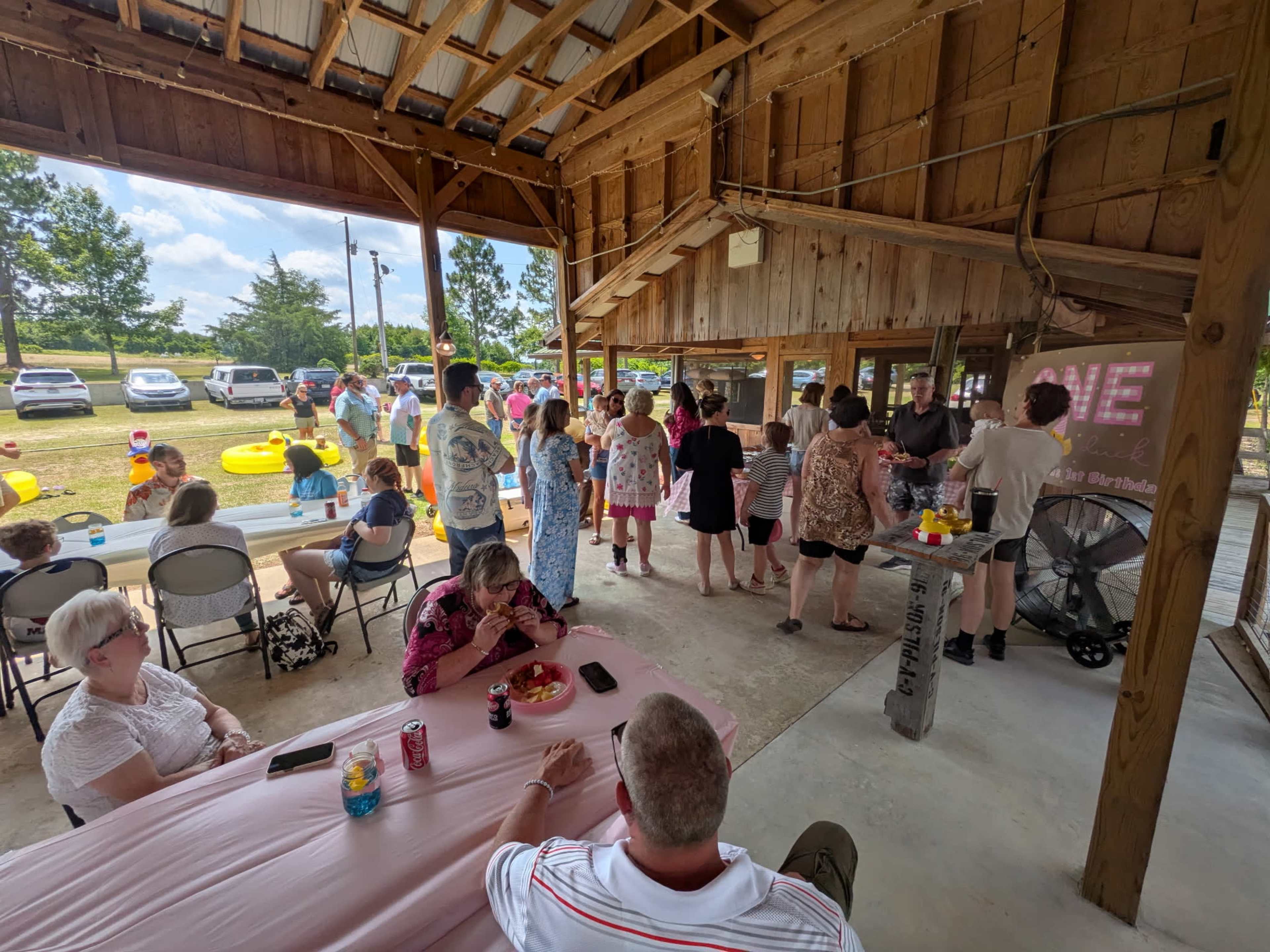 A gathering of people mingles under a wooden pavilion during an outdoor event, with tables set up and decorations visible in the background.
