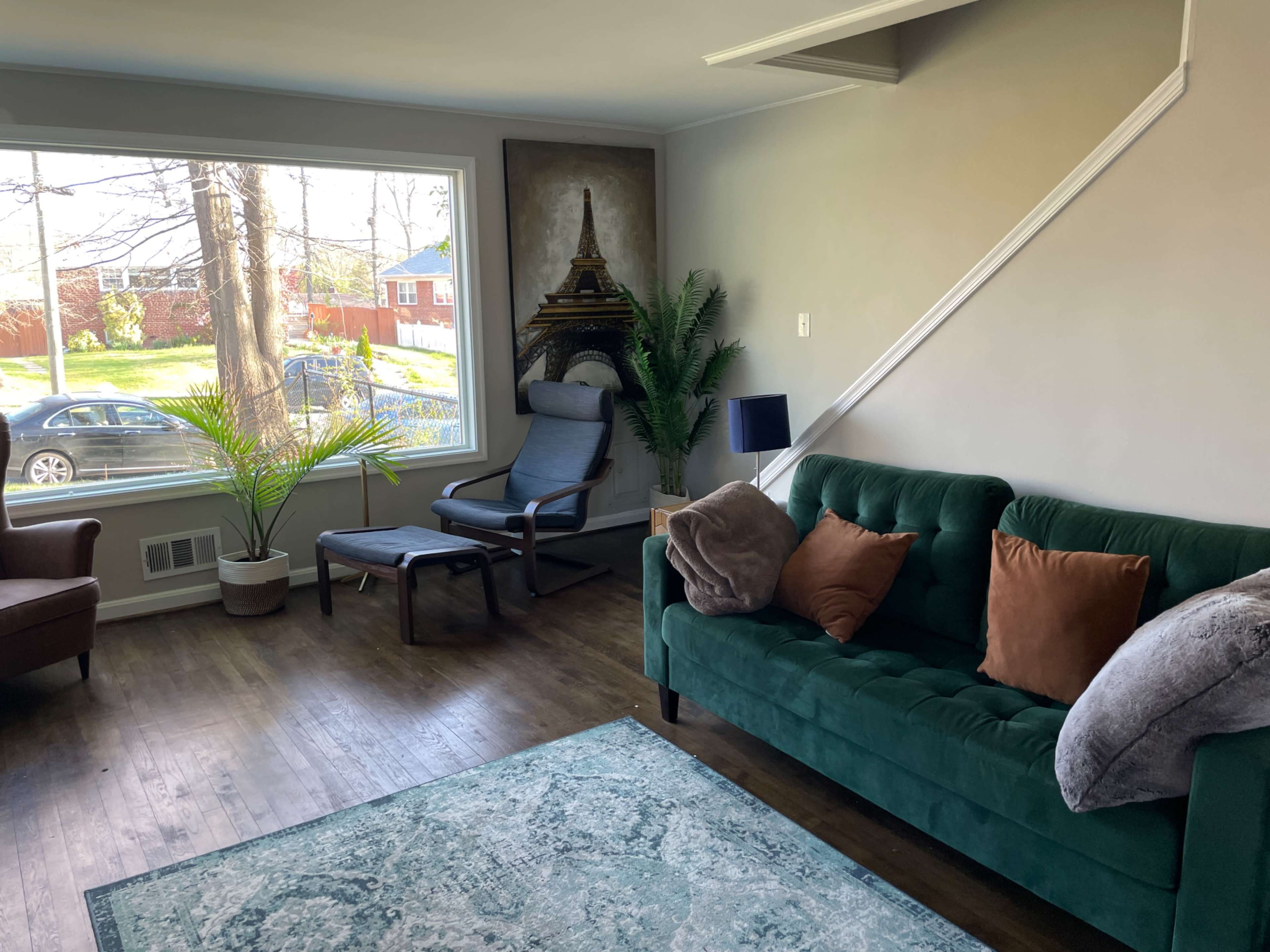 A living room features a green sofa with brown pillows, a blue chair, a large window with a view of the outdoors, and a decorative rug on the wooden floor.