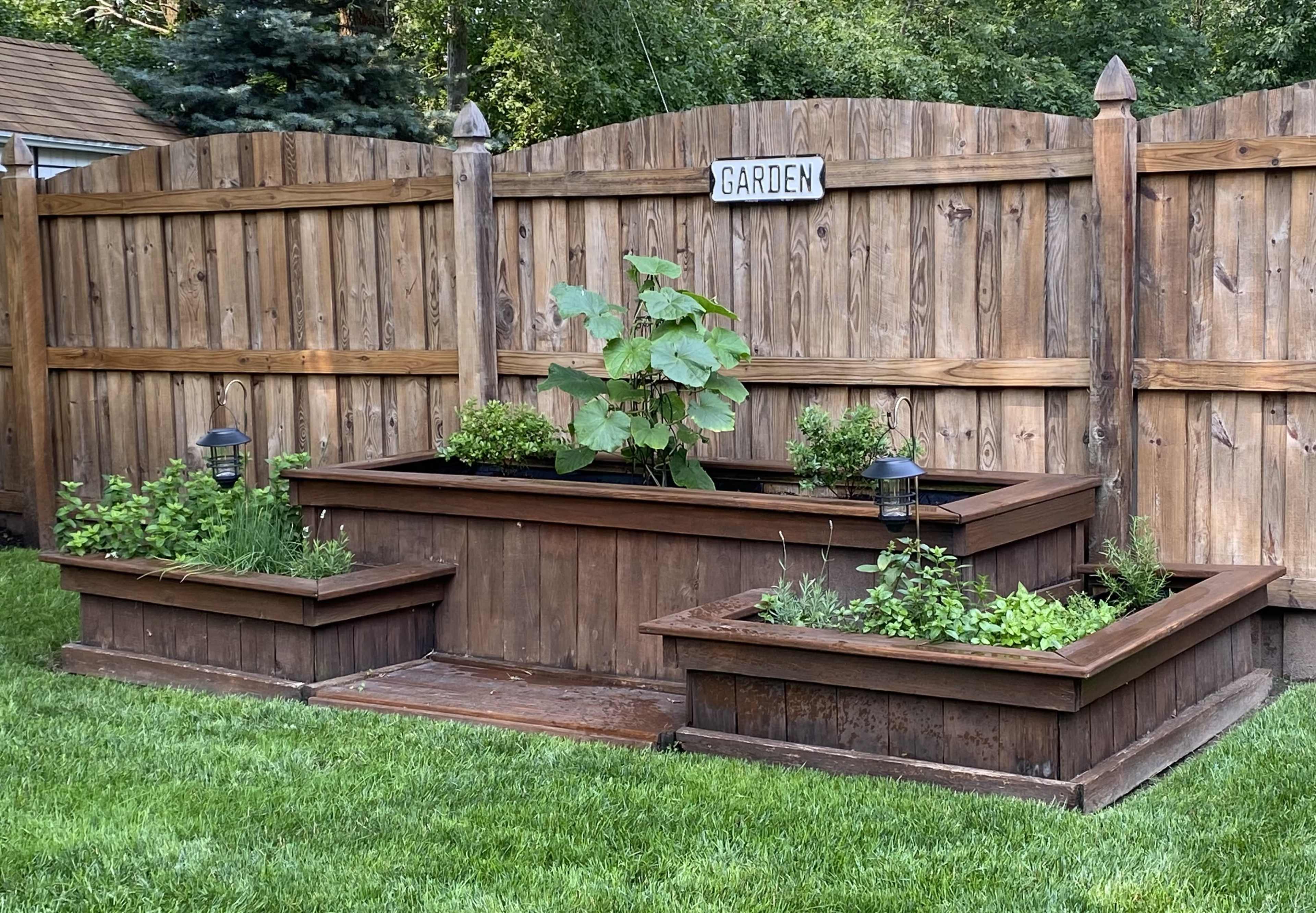A wooden garden bed with a flowering plant in the center, surrounded by smaller raised planters, all set against a wooden fence.