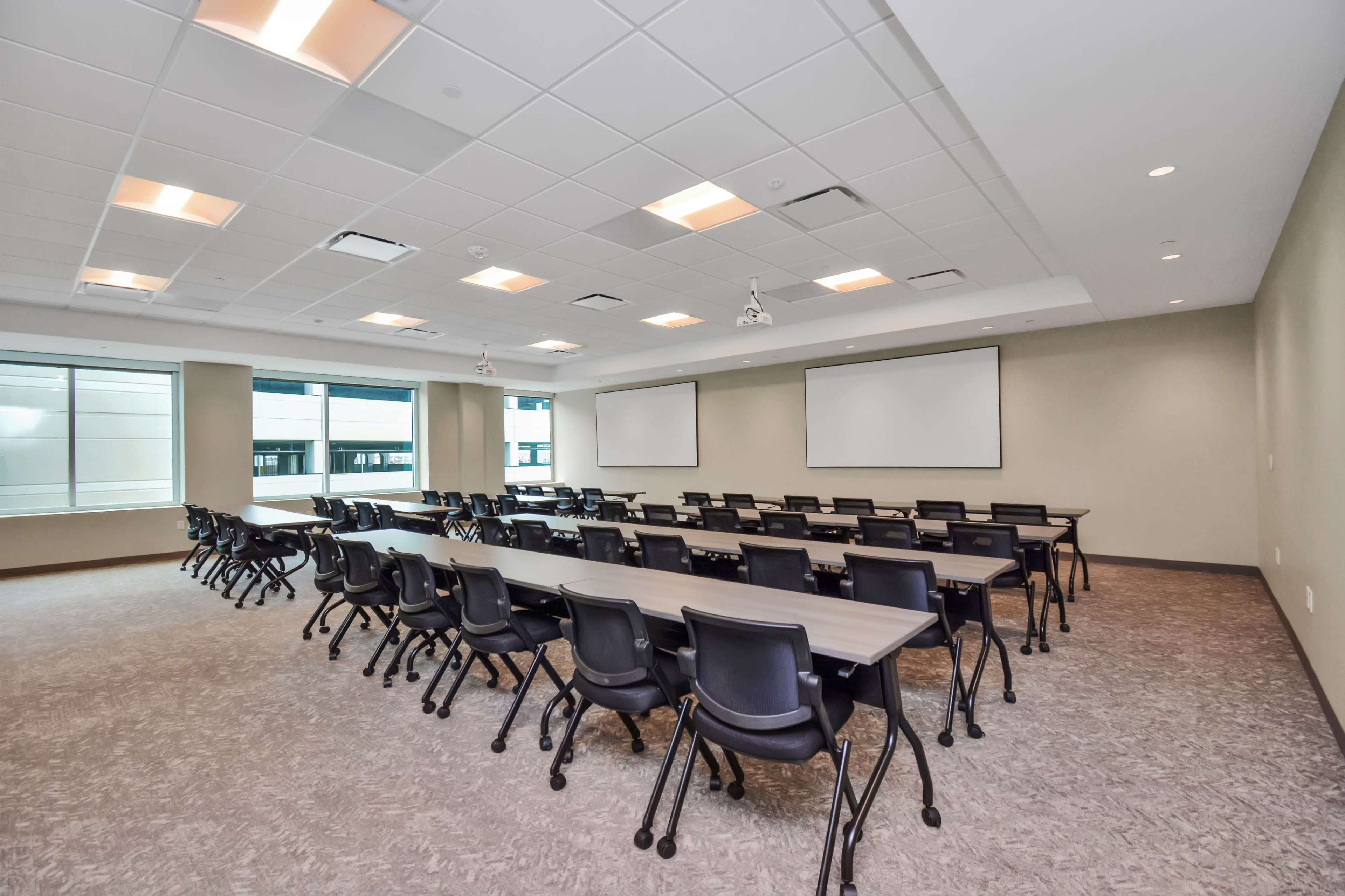 A modern conference room features several rows of black chairs facing two blank projection screens against a light-colored wall.