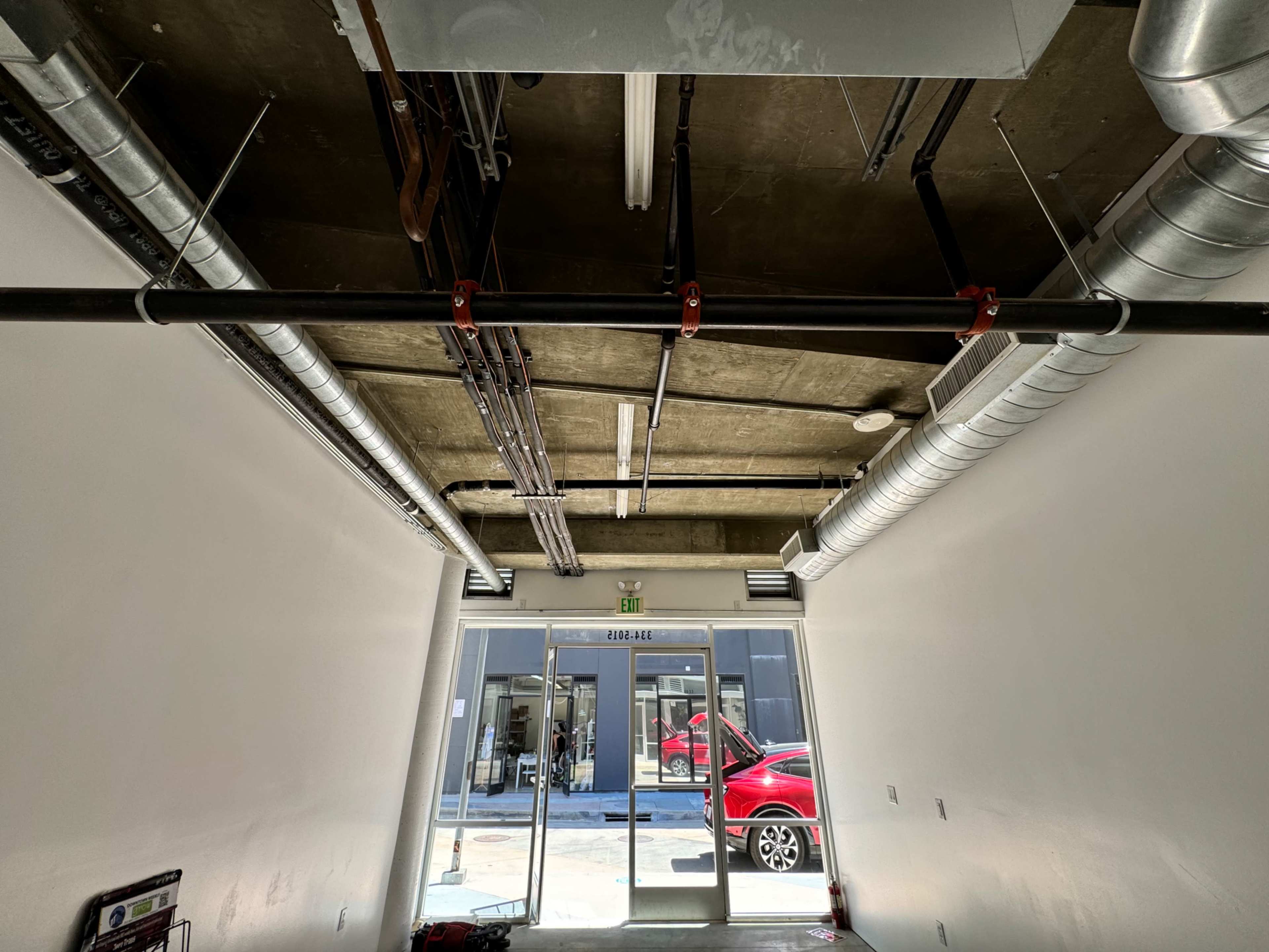 An industrial-style hallway with exposed metal pipes and a view of a red car parked outside through glass doors.