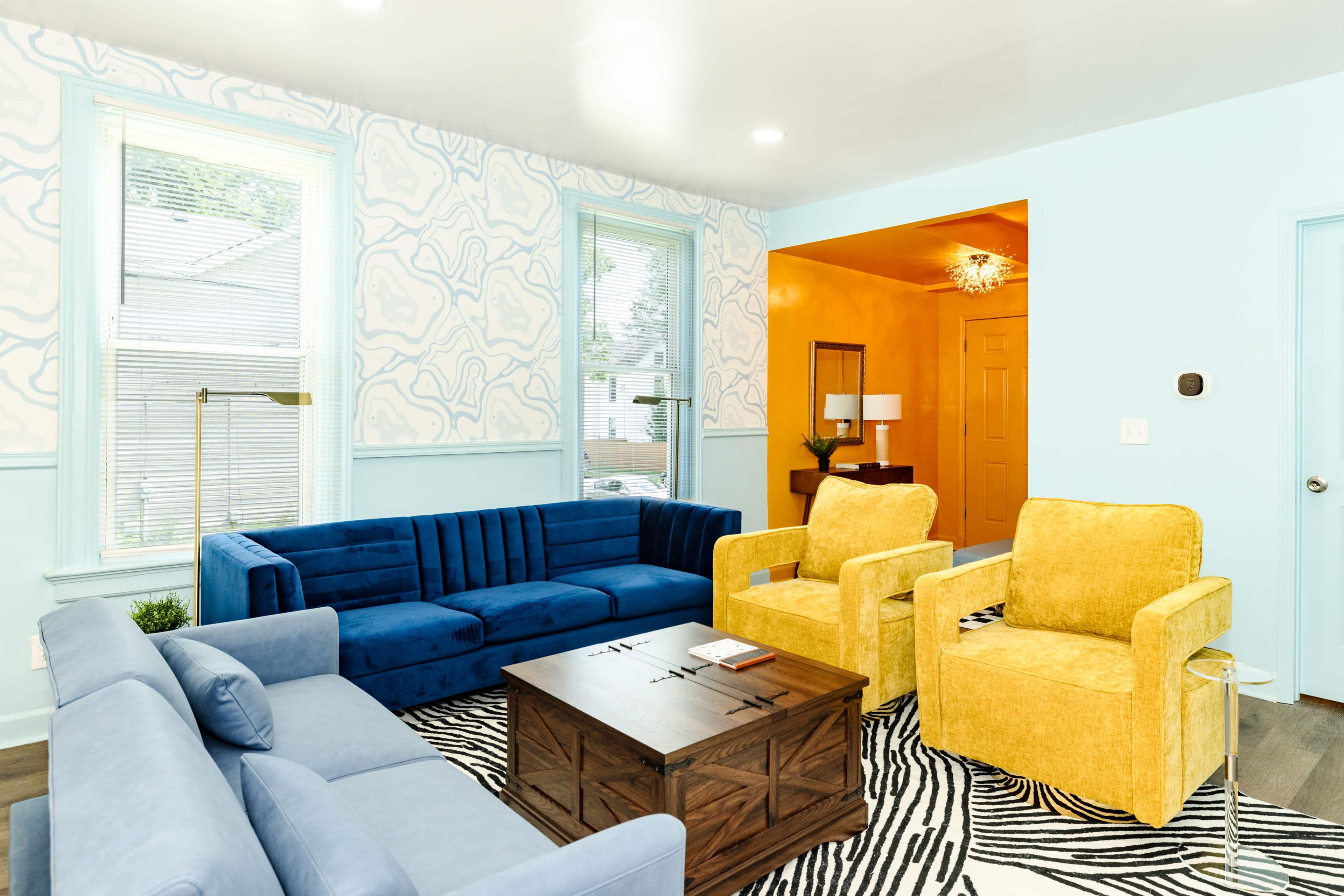 The living room features a blue sofa, yellow armchairs, and a wooden coffee table on a zebra-striped rug, with light blue walls and large windows.
