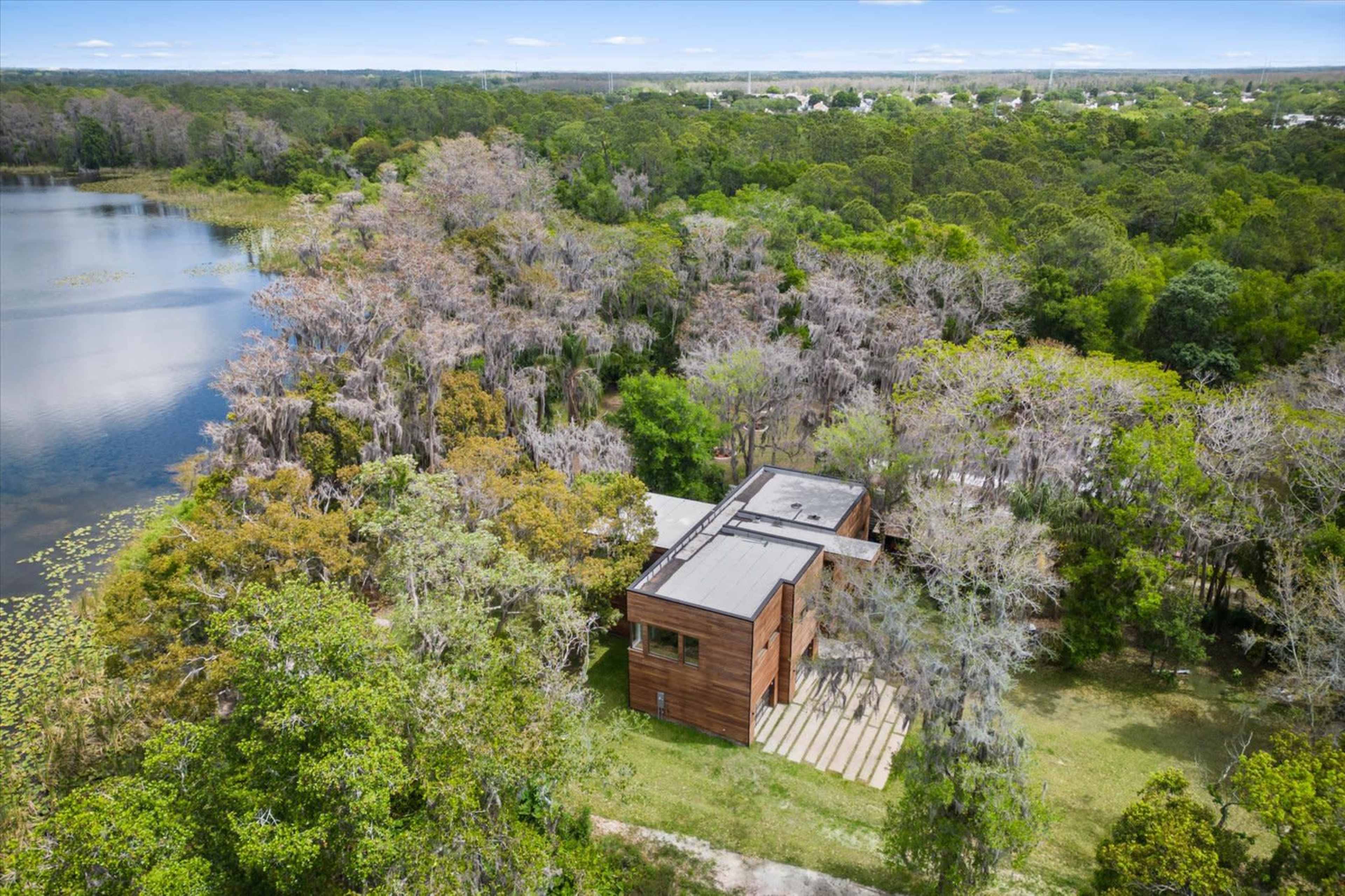 The image shows a modern wooden house situated near a body of water, surrounded by trees and greenery.