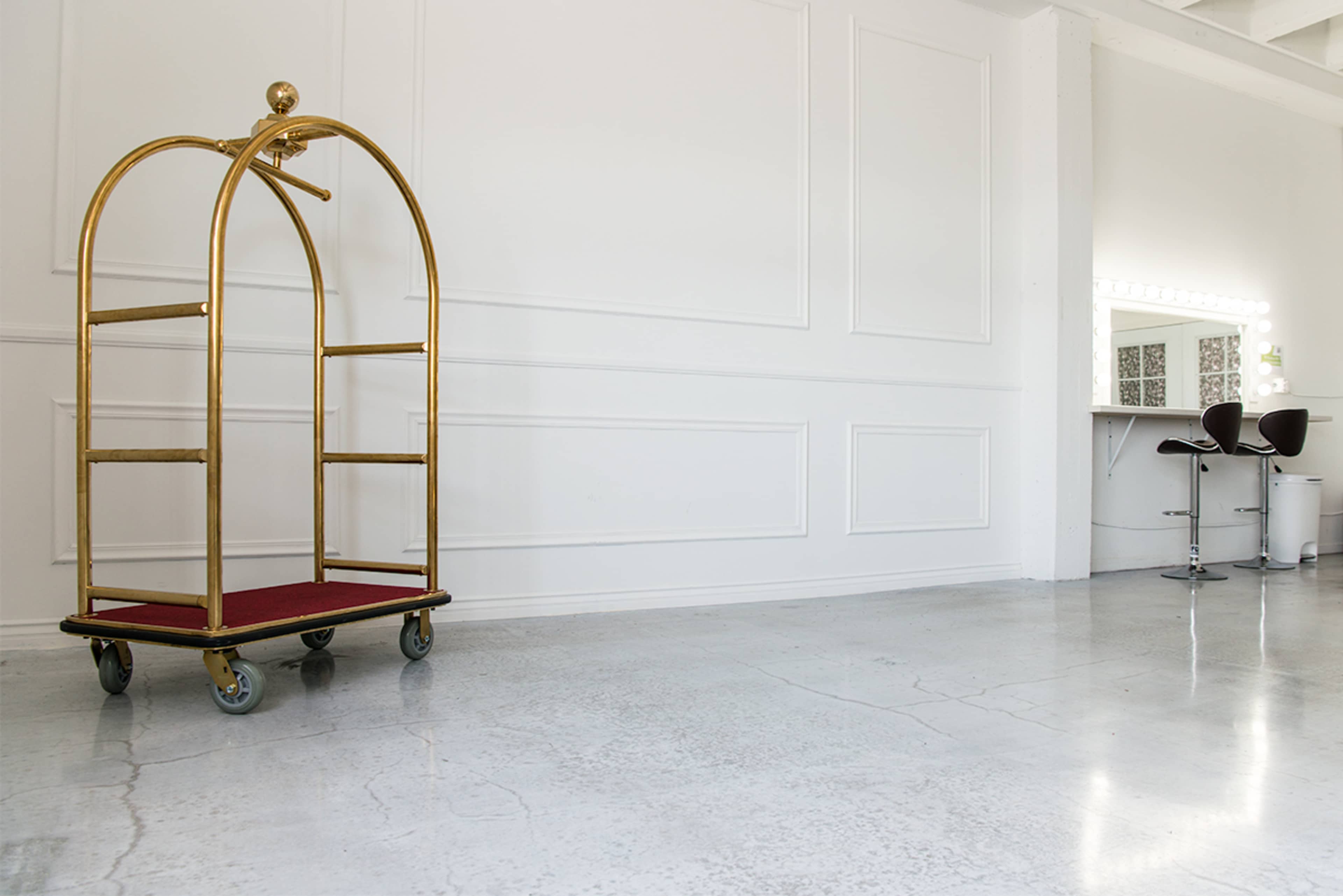 A brass luggage cart on wheels placed against a white wall, with a mirrored vanity and two black chairs visible in the background.