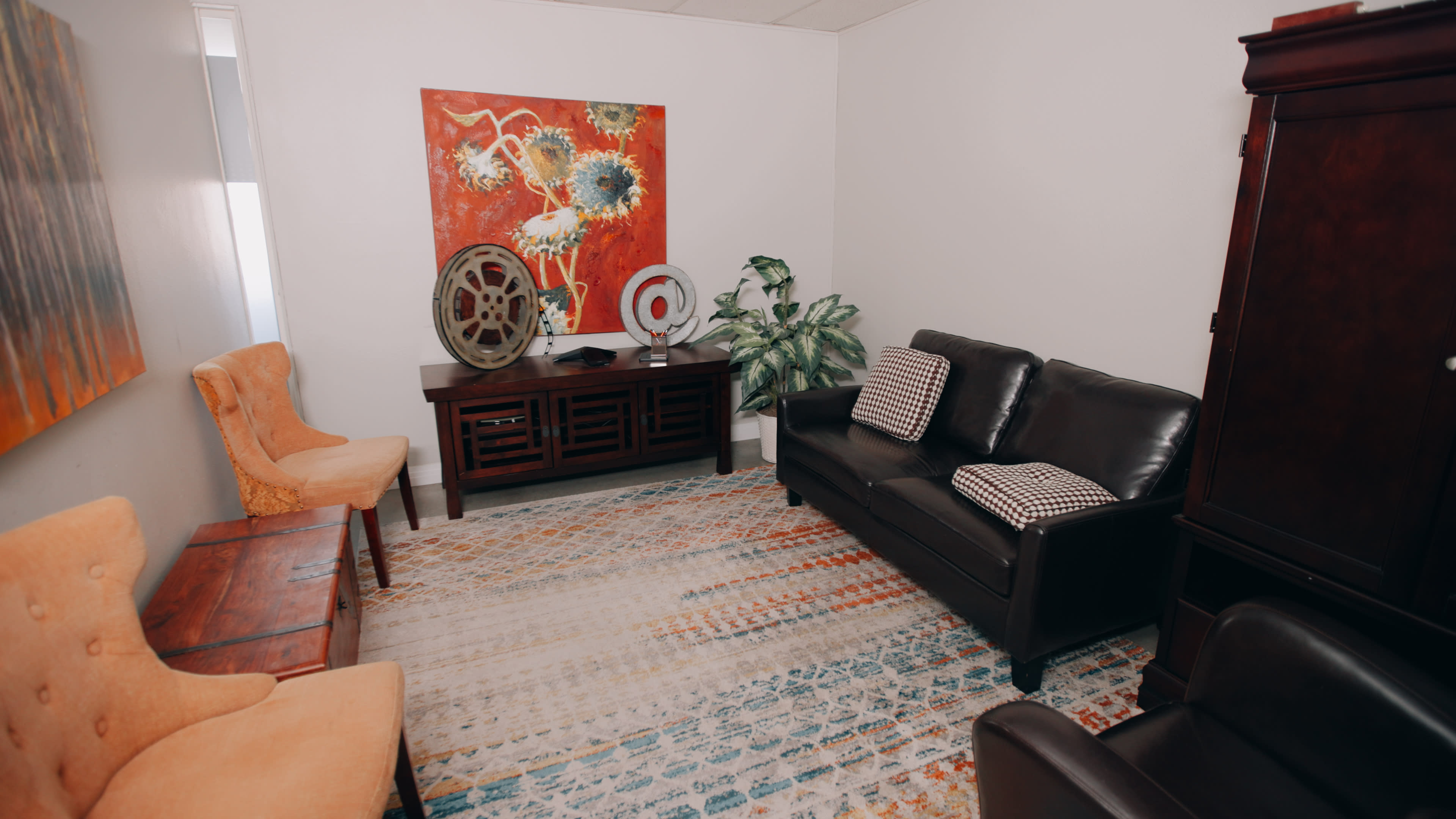 A small, furnished room with a black sofa, two orange chairs, a wooden table, a colorful rug, and a large decorative painting on the wall.