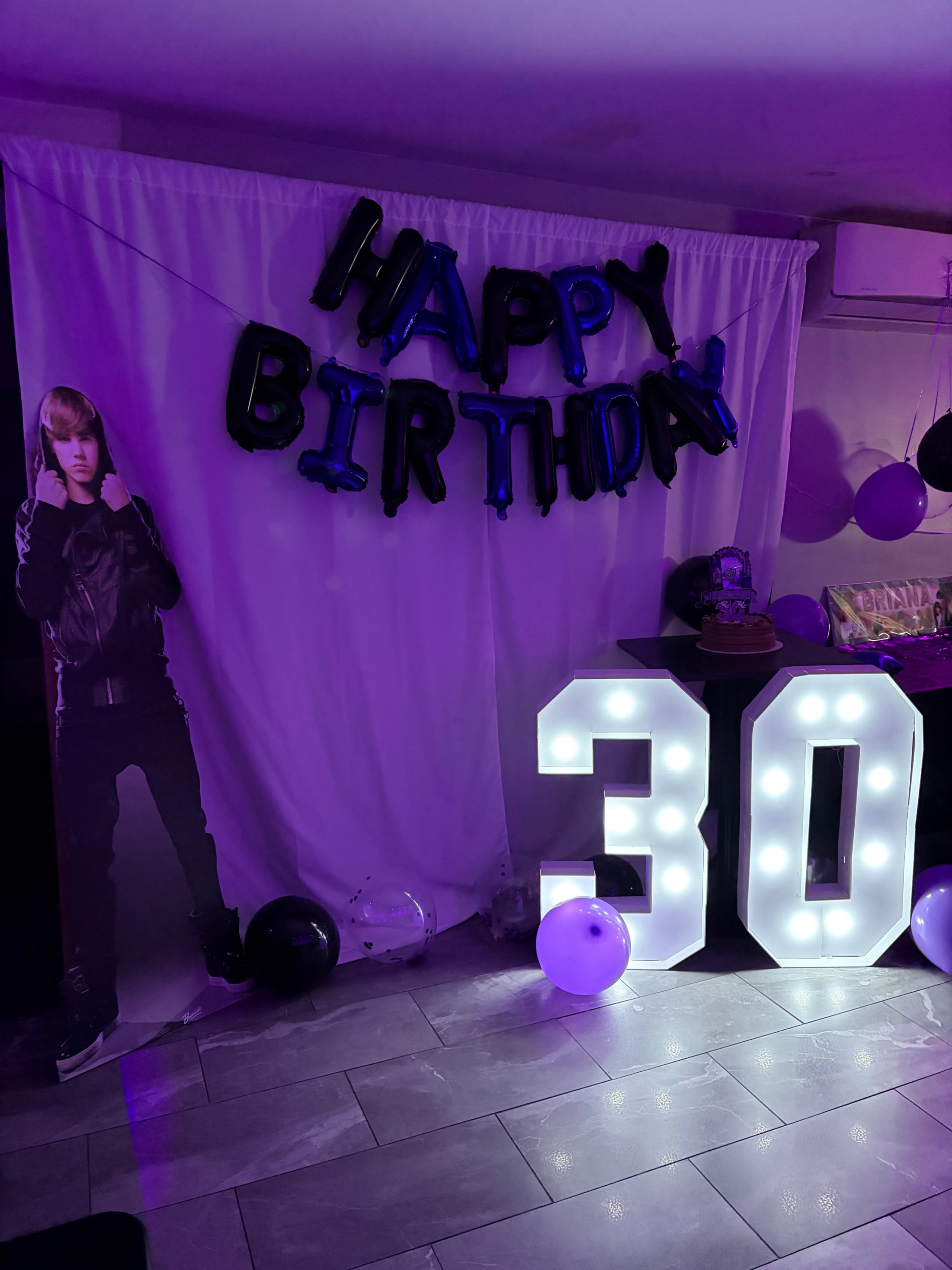 The scene features a birthday celebration backdrop with a "Happy Birthday" banner, a life-sized cutout of a person, and illuminated "30" numerals surrounded by black and purple balloons.