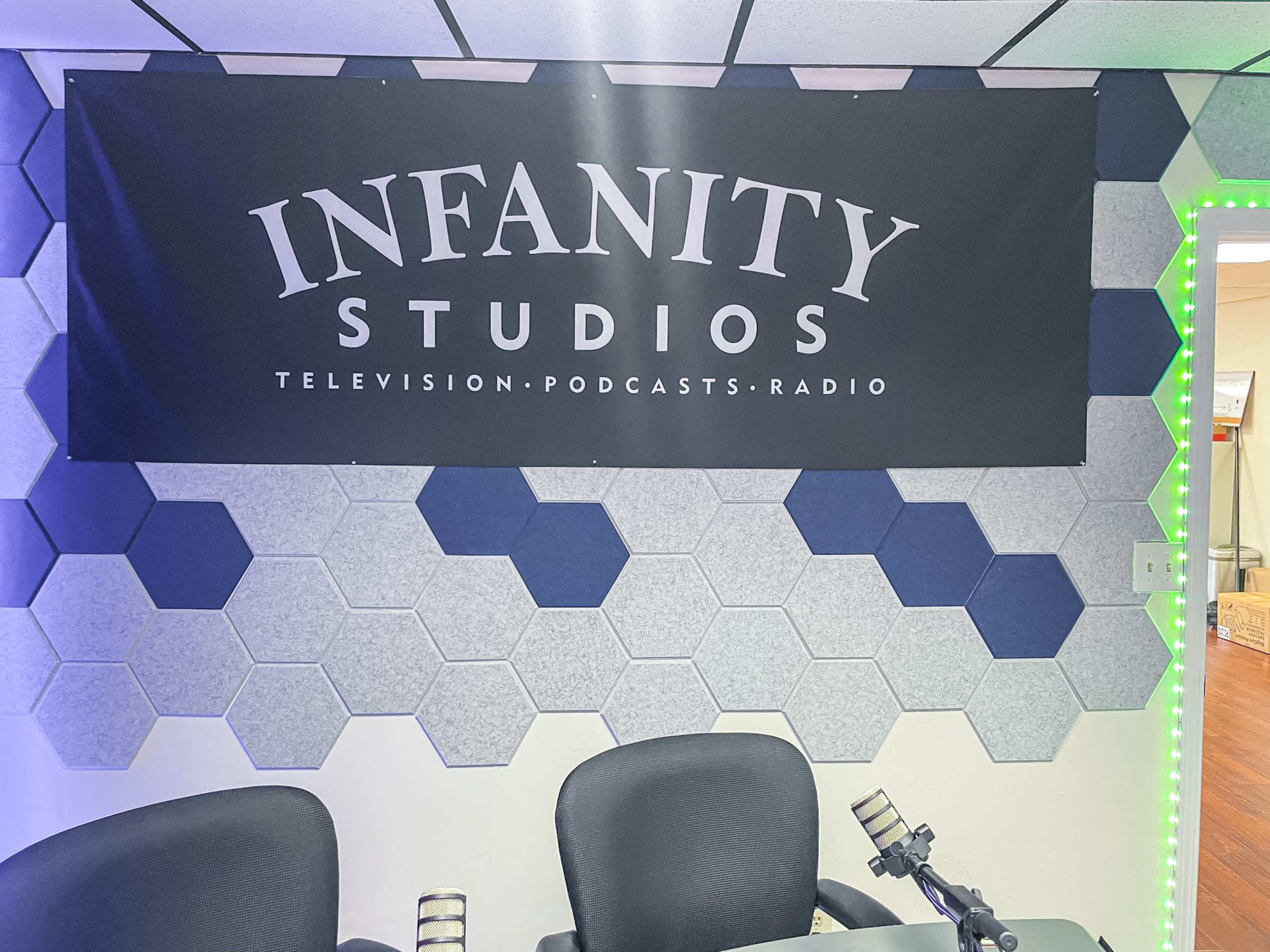 A wall adorned with hexagonal sound panels, featuring a banner that reads "INFINITY STUDIOS" along with the text "TELEVISION. PODCASTS. RADIO." in a podcast studio setting.