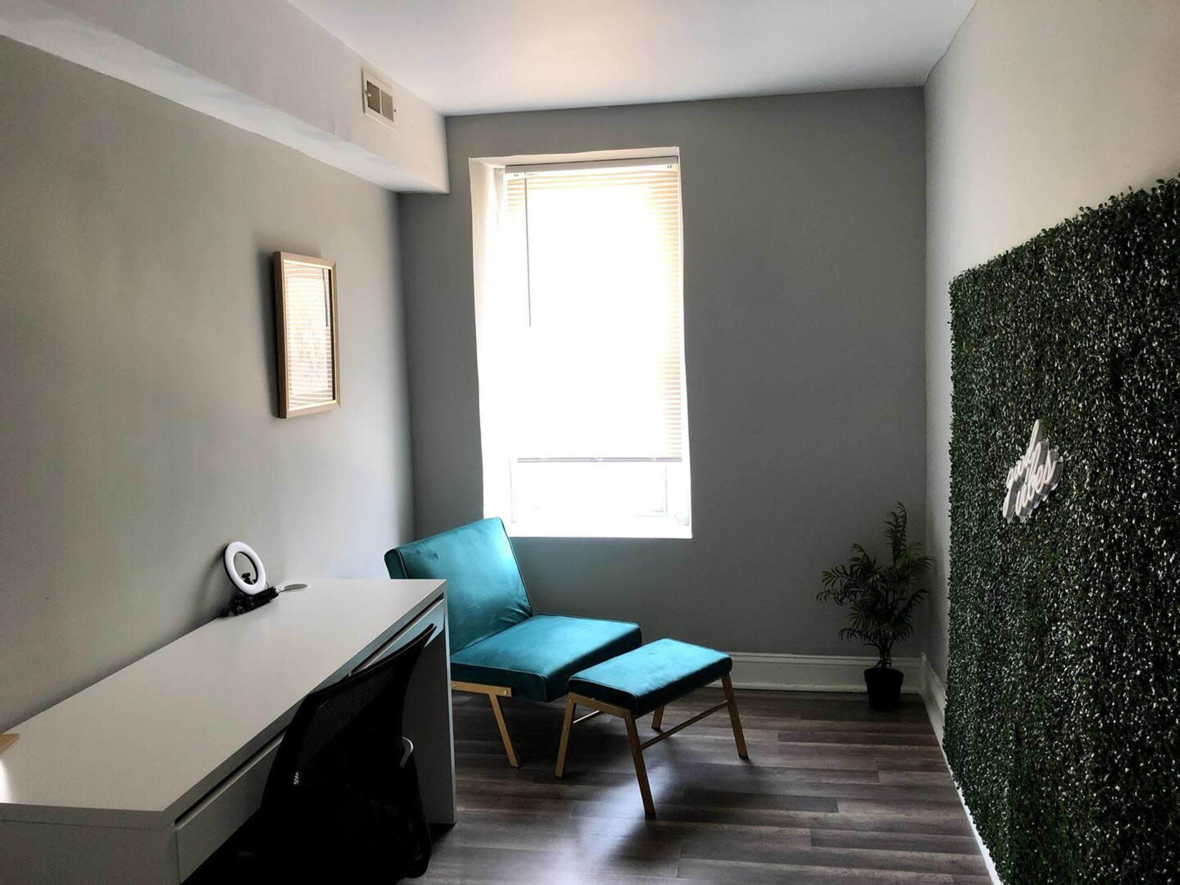 A small, minimalist room with a teal chair and ottoman, a white desk with a black chair, and a wall covered in green foliage near a window.