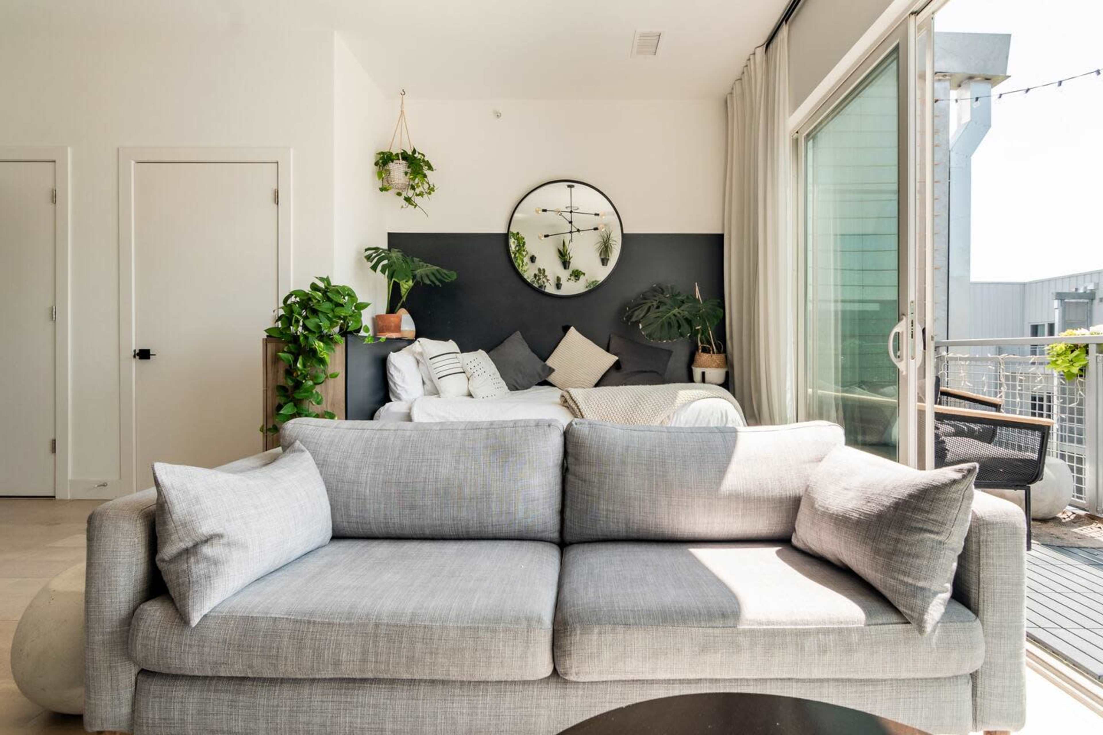 A modern living space features a gray sofa, a round wall clock, potted plants, and a bed visible in the background.