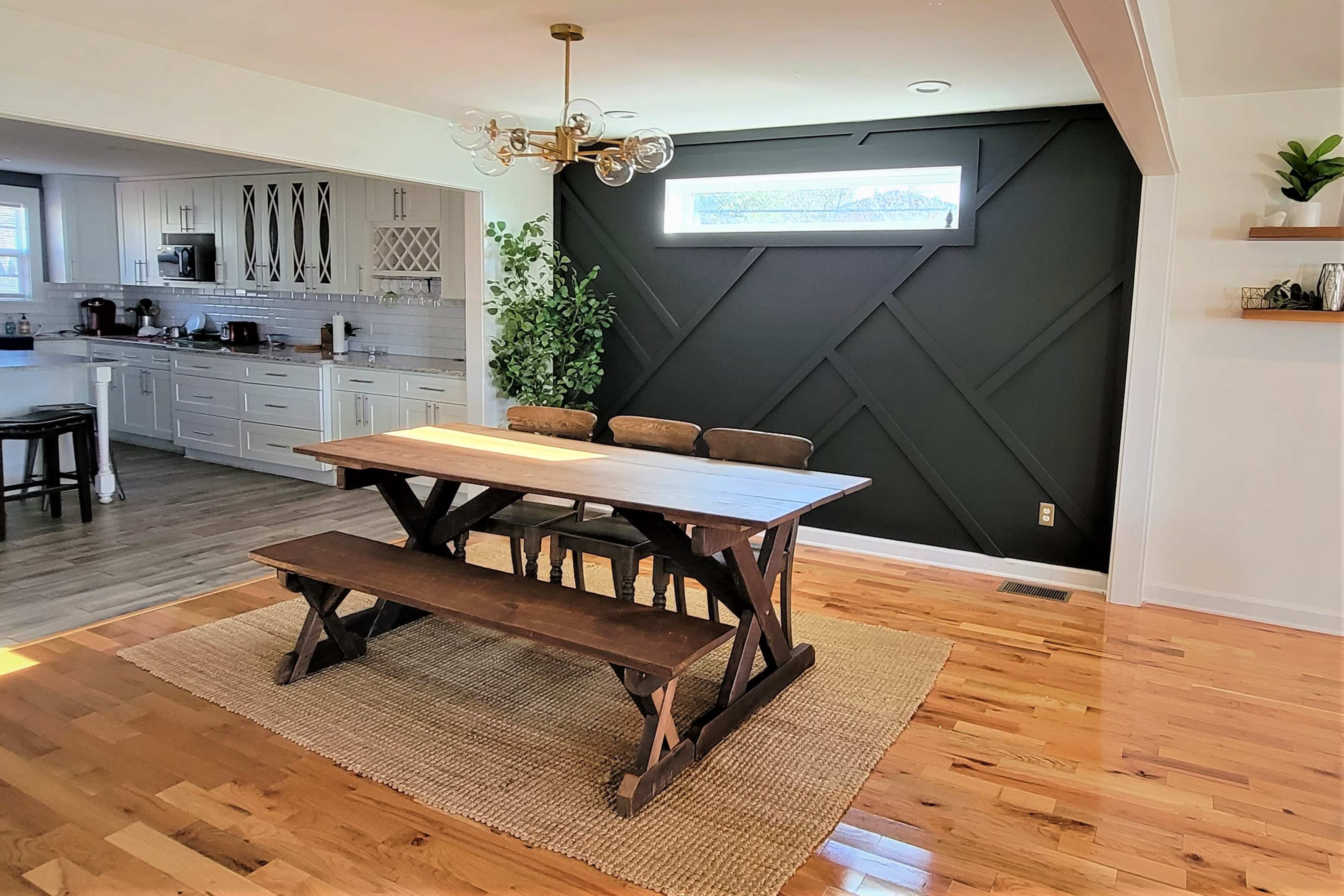 A wooden dining table with matching benches is set on a woven rug in a brightly lit dining area featuring a black accent wall and a view into a kitchen.