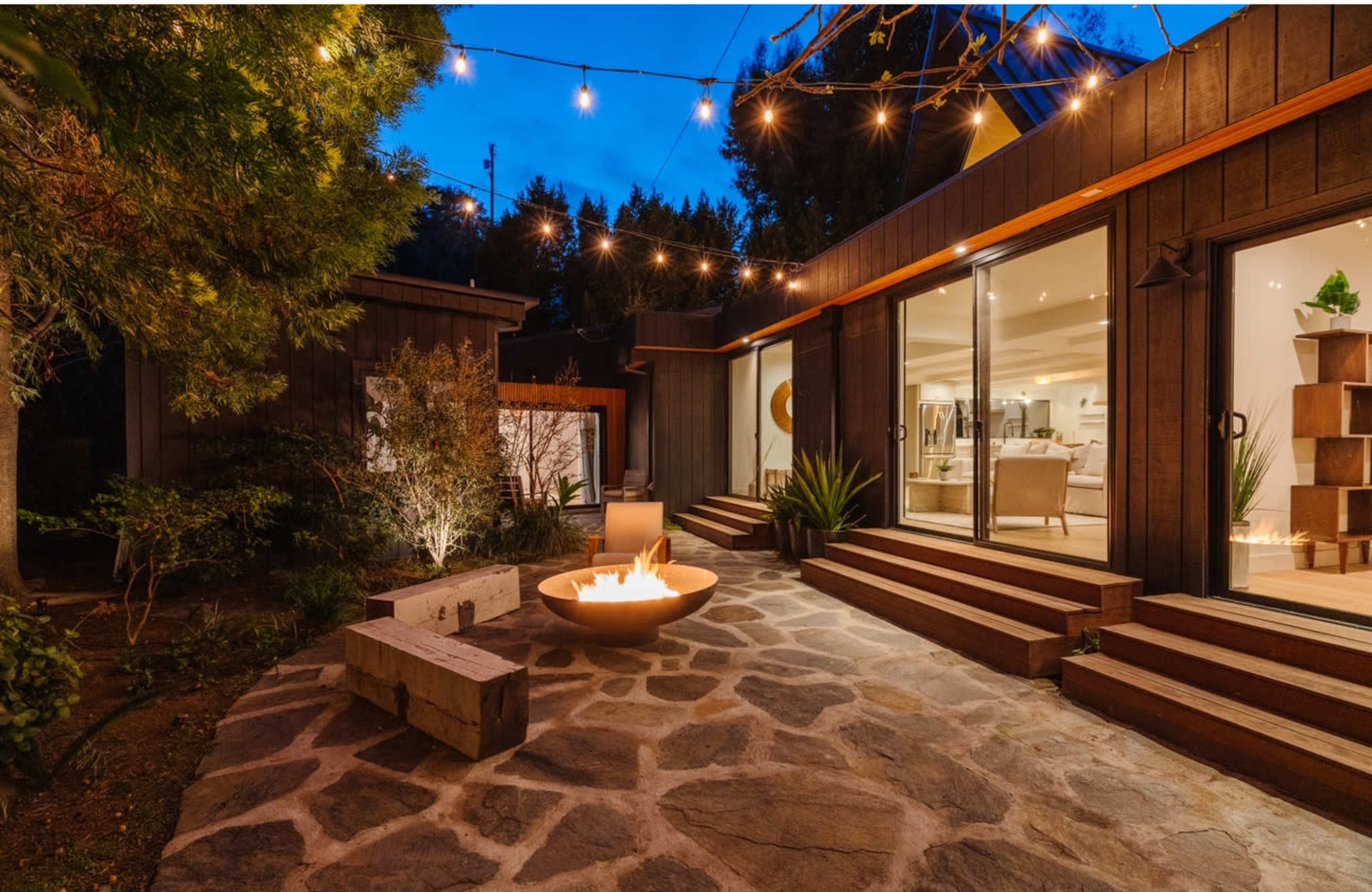 A stone patio features a fire pit surrounded by steps leading to a contemporary wooden structure, illuminated by string lights at dusk.