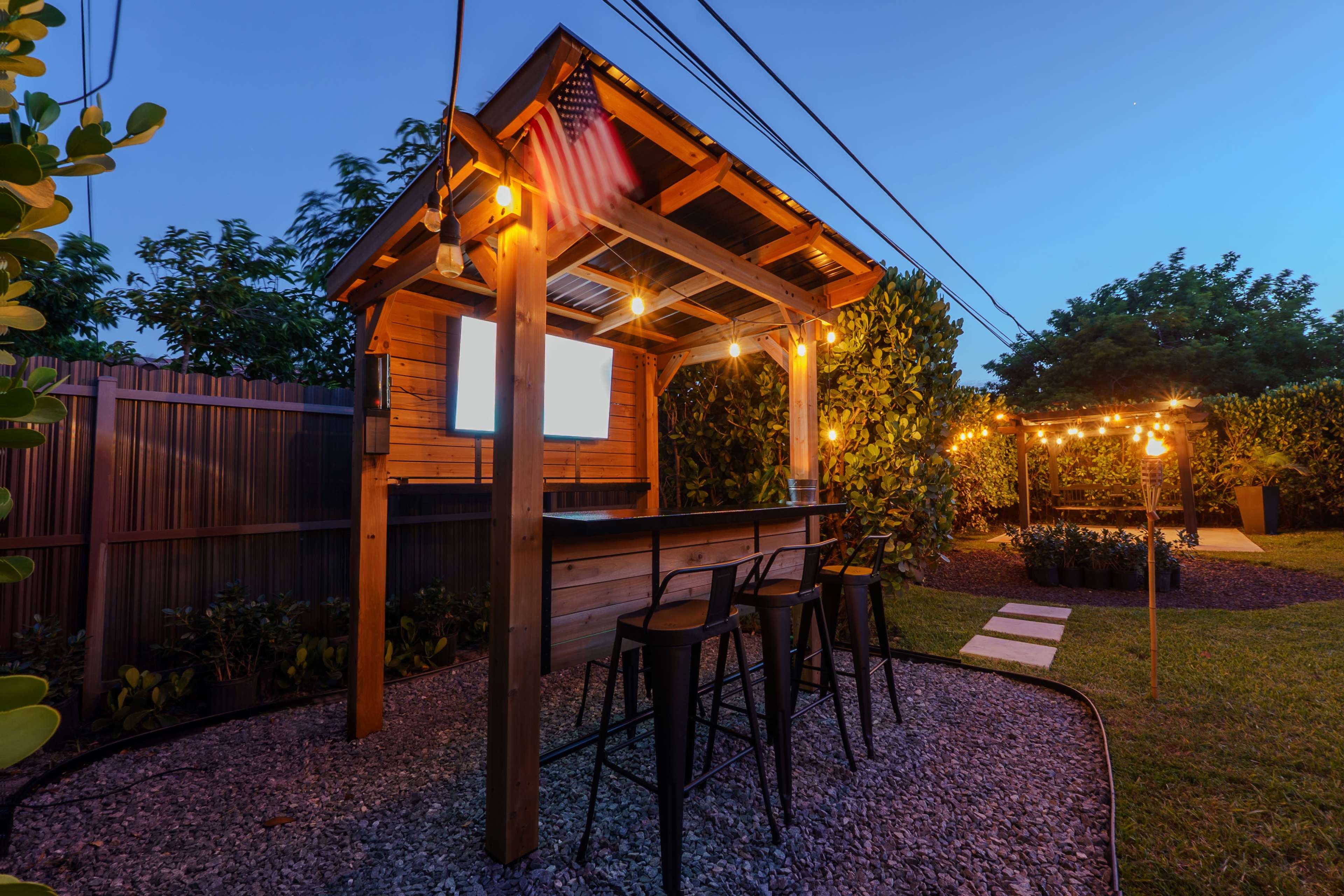 A wooden outdoor bar with high stools is illuminated by string lights in a garden setting at dusk.