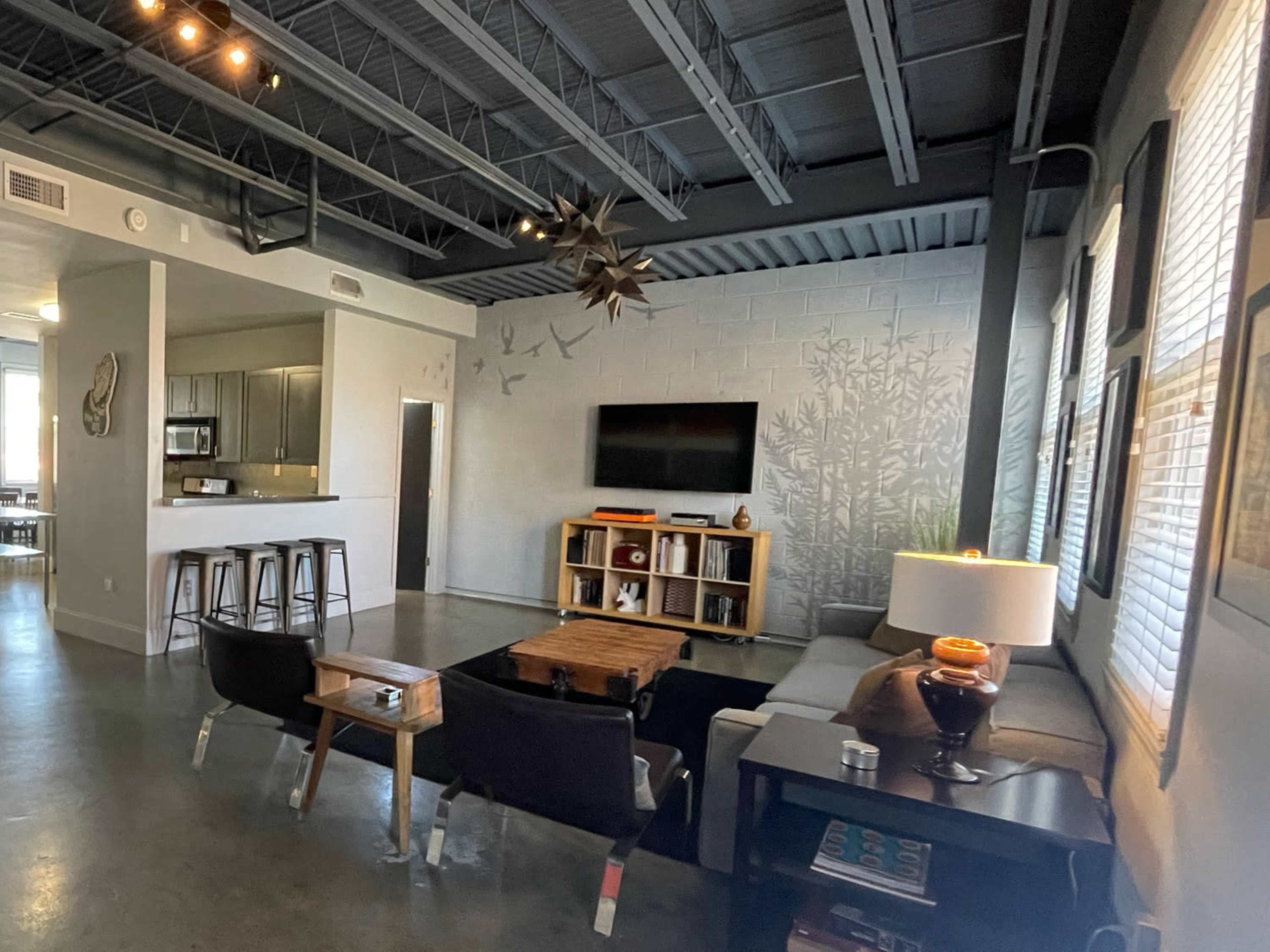 The image shows a modern living room with a concrete floor, a sectional couch, a wooden coffee table, and a wall-mounted television opposite a kitchen area.