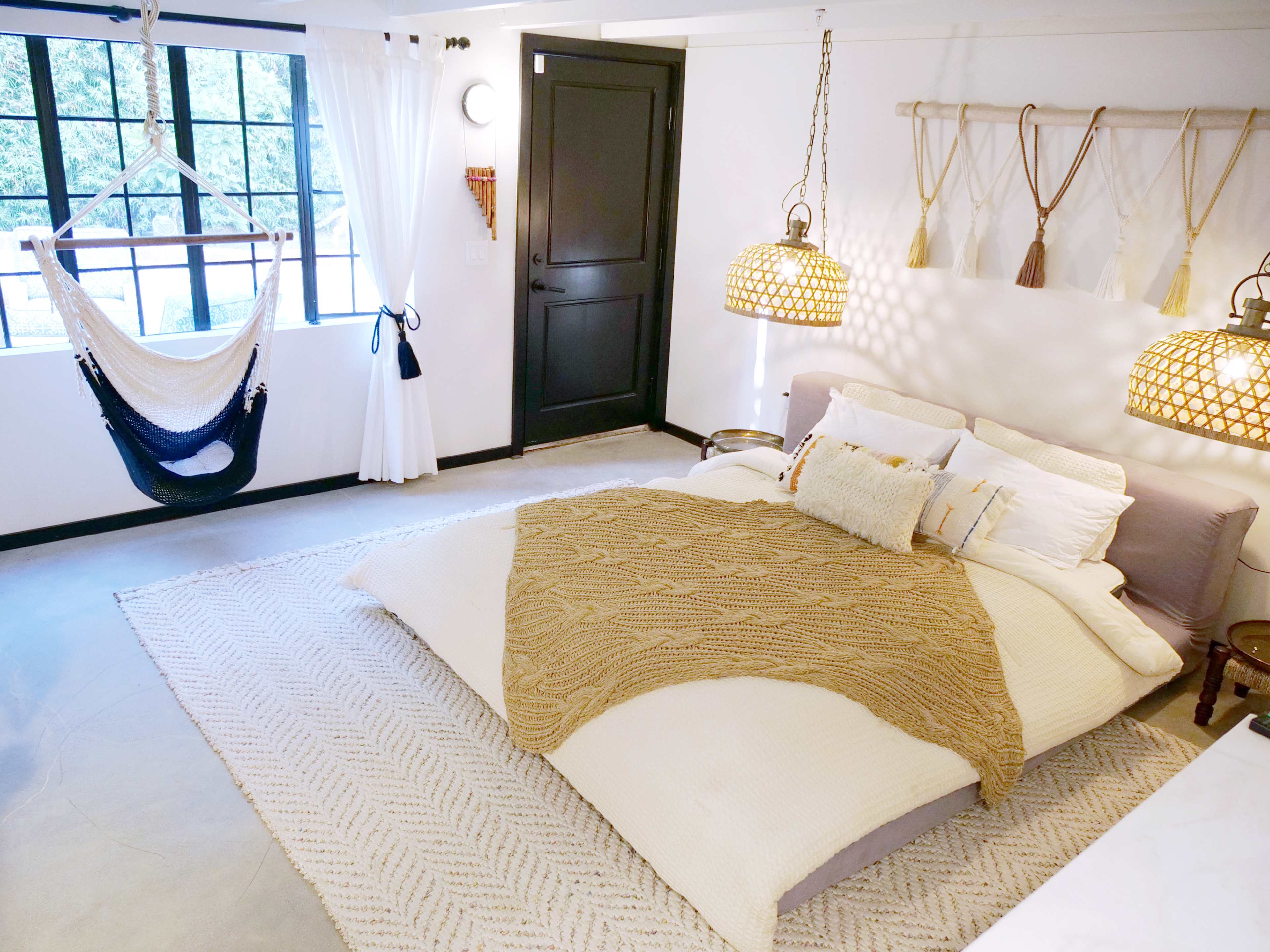 A well-lit bedroom features a large bed with textured bedding, a hanging hammock, and woven light fixtures against minimalist decor.