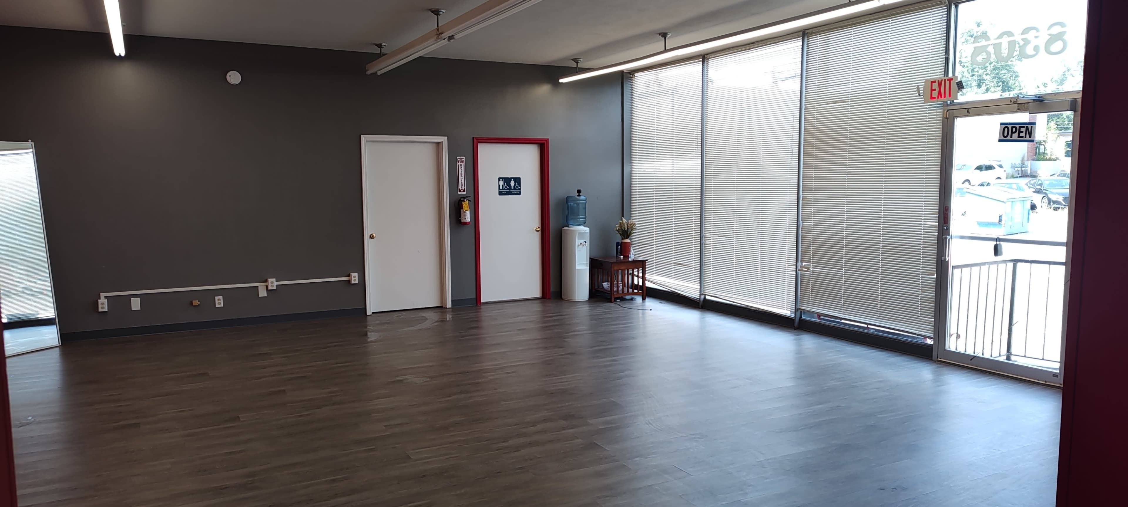 The image depicts an empty interior space with gray walls, a wooden floor, a single entrance door, a water cooler, and large windows letting in natural light.