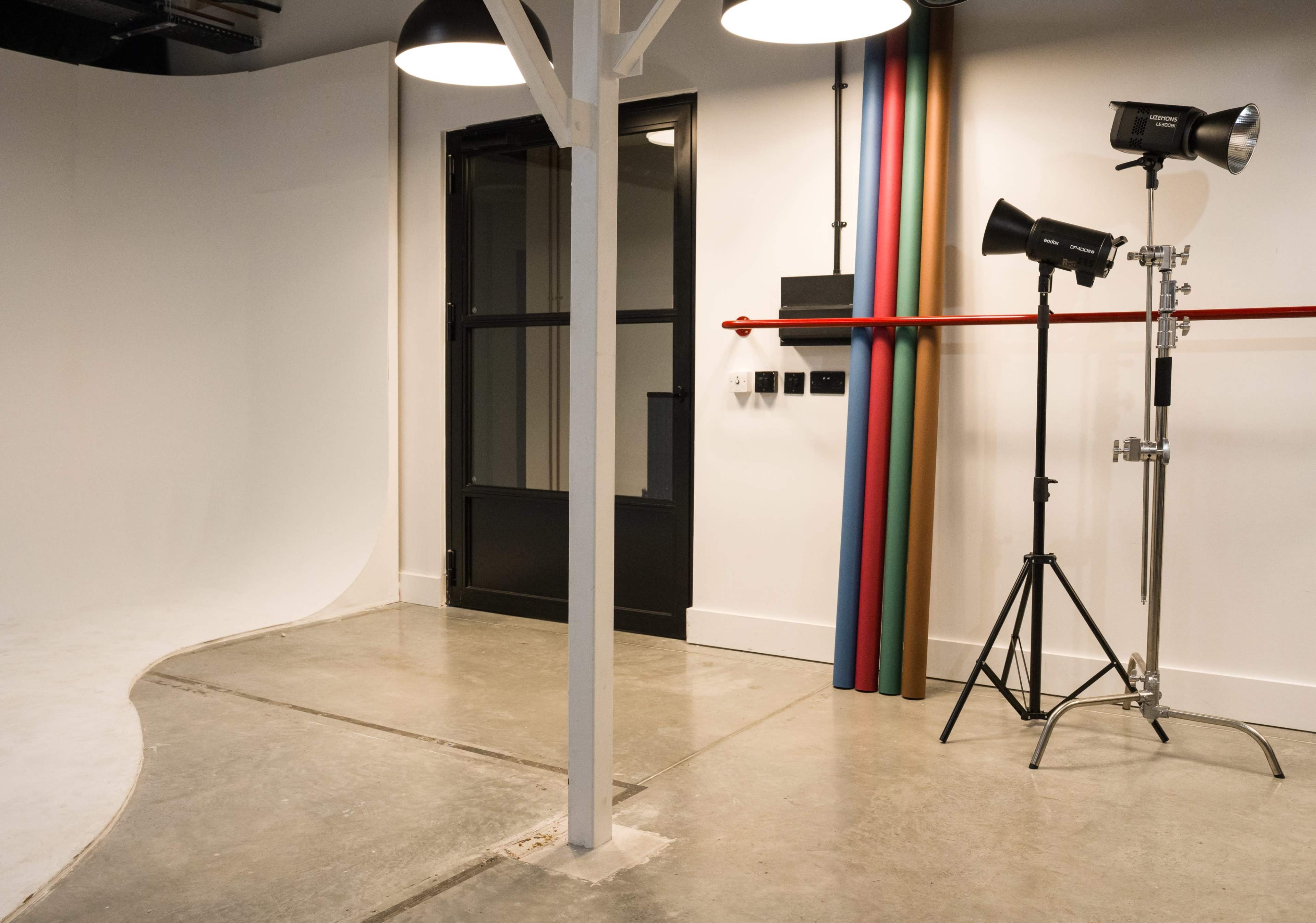 The image shows a photography studio with a curved seamless backdrop, a black door, and two light stands positioned next to colorful backdrop rolls.