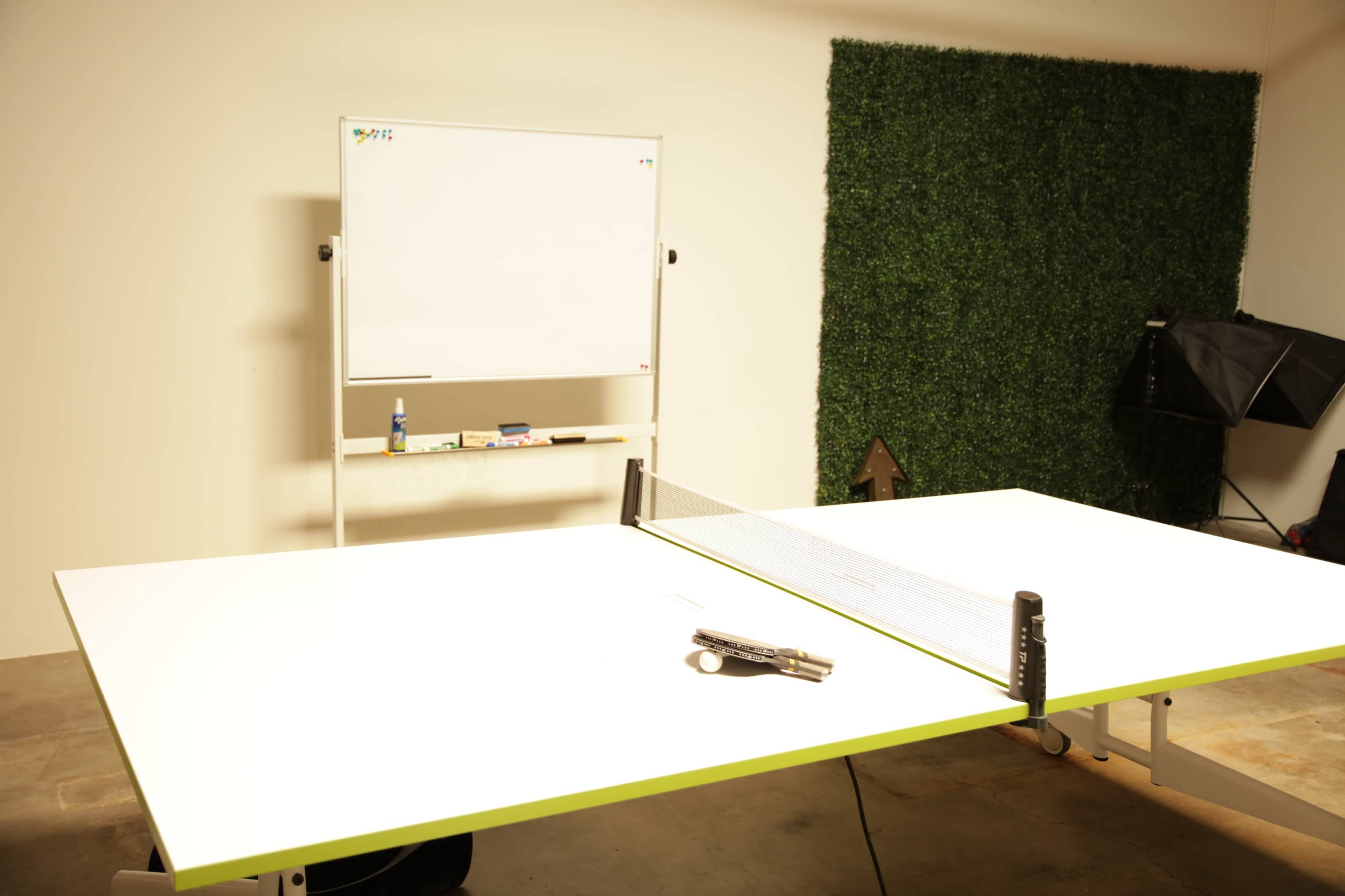 A large, white table with a tennis net setup is positioned in front of a whiteboard and a green wall, with a tool lying on the table.