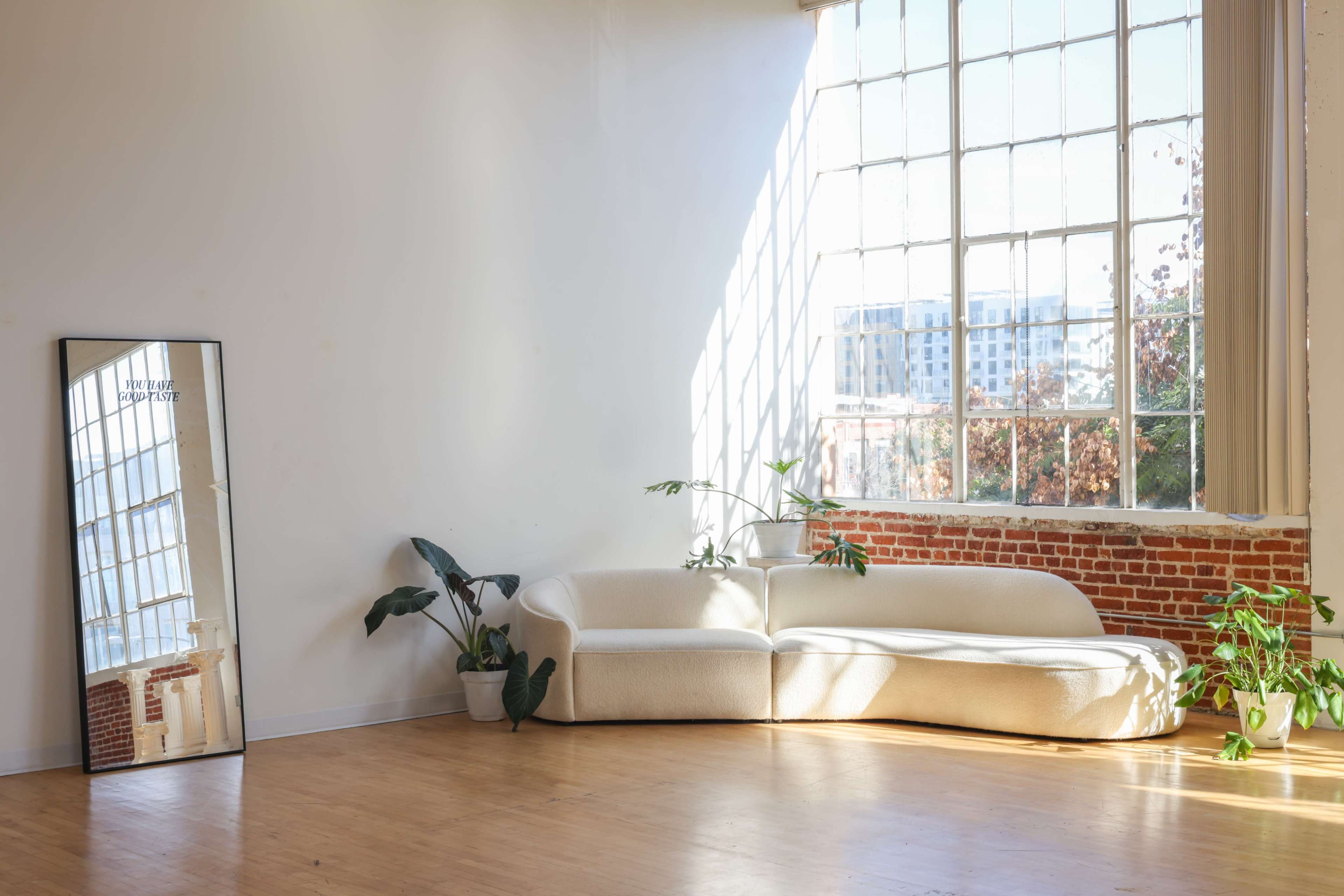 A curved white sofa is positioned next to a large window in a bright, minimalist room with wooden flooring and potted plants.