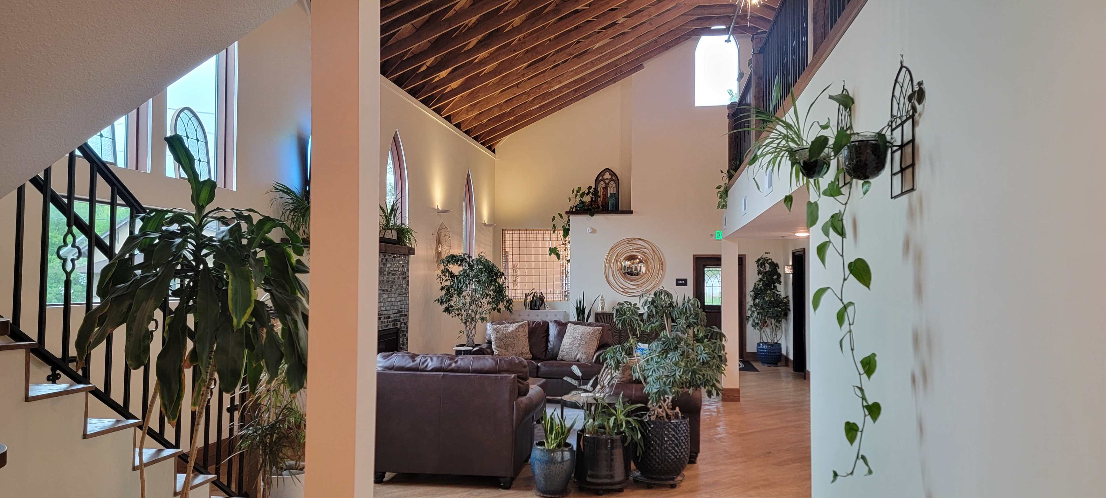 The image shows a spacious living area with high ceilings, large windows, and various plants arranged around a seating area and staircase.