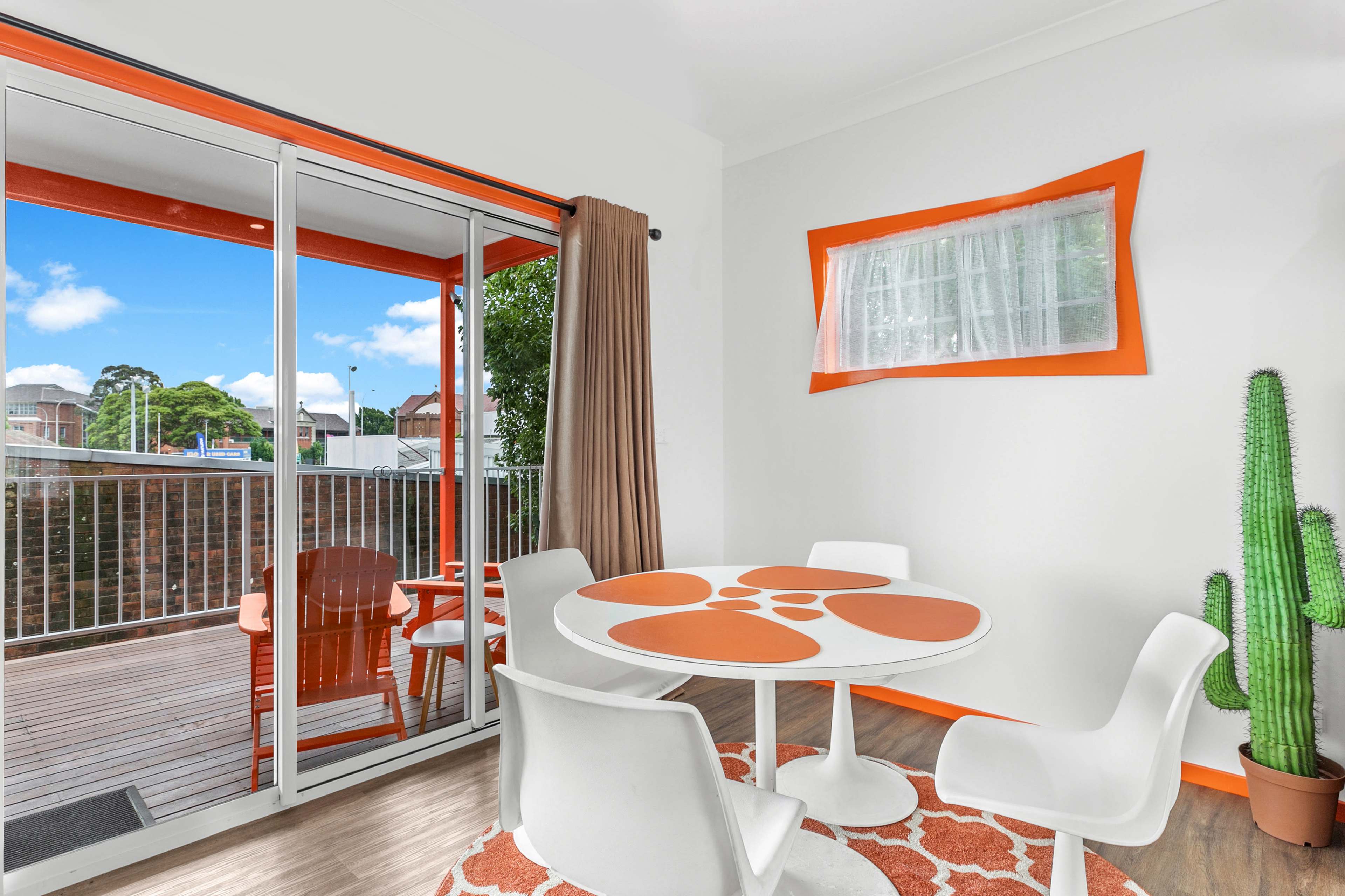 A dining space featuring a circular table with orange accents, surrounded by white chairs, and a view of a balcony through sliding glass doors.