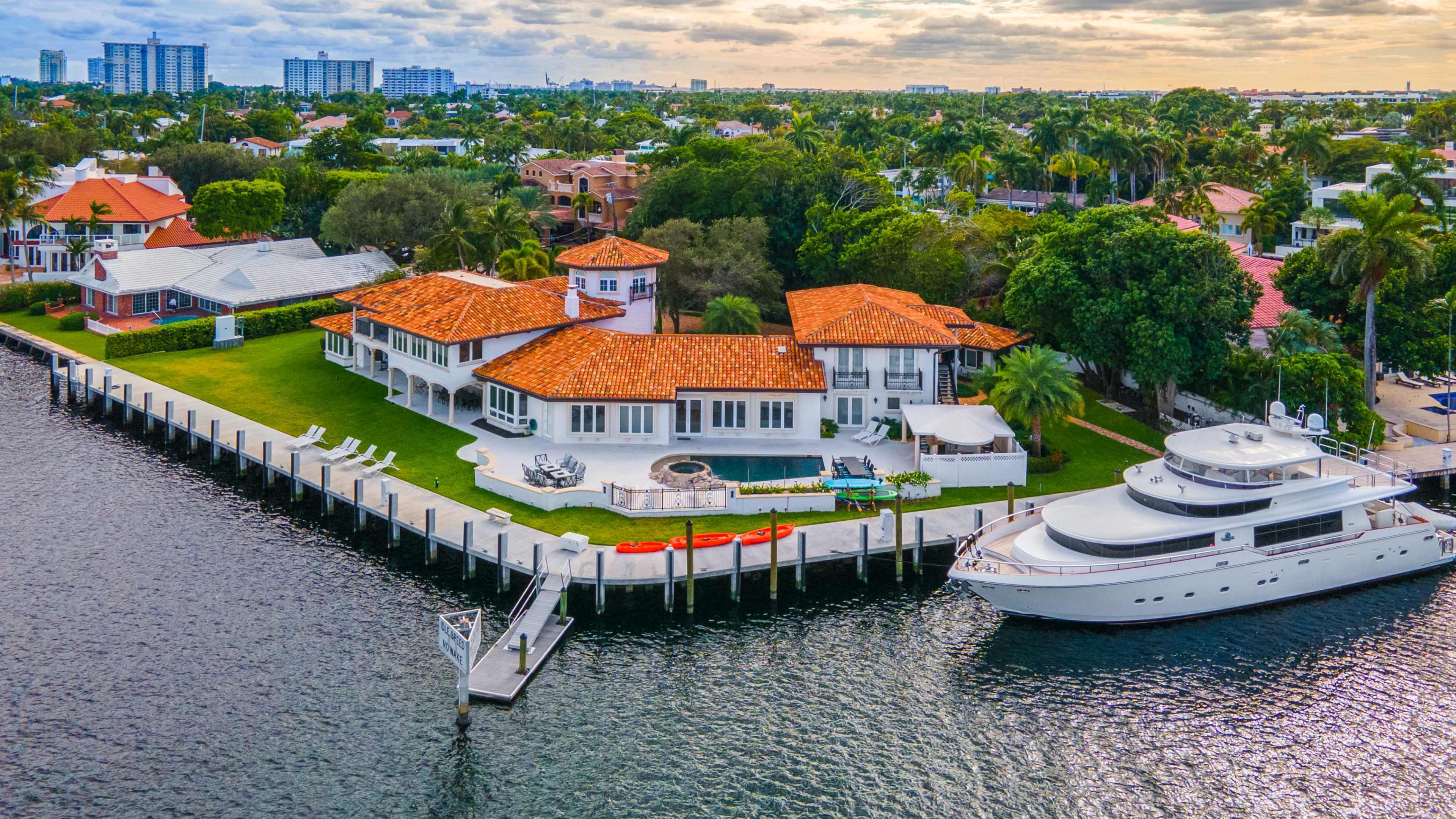 The image shows a waterfront luxury home with a large lawn, a swimming pool, and a docked yacht.
